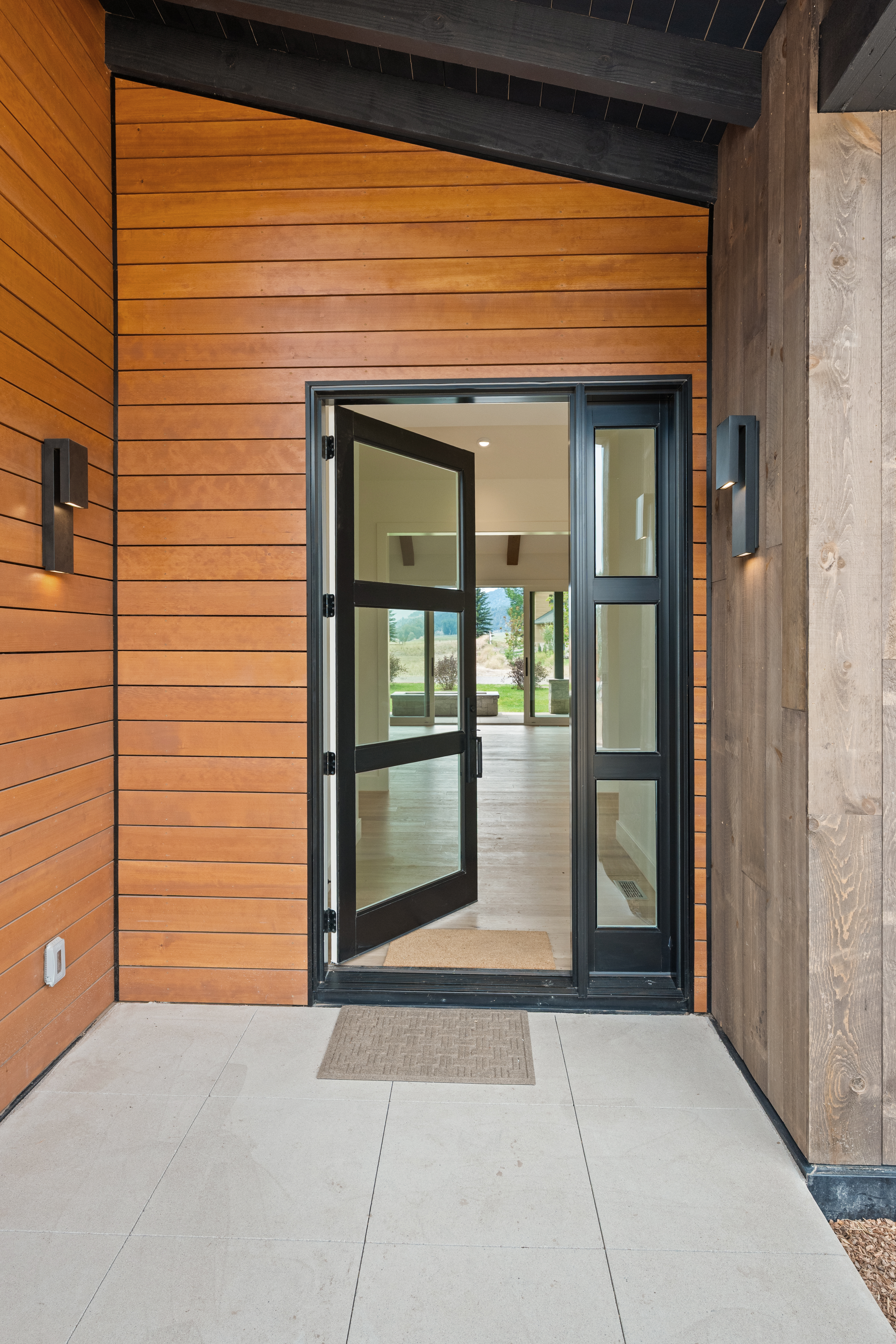 This modern, architectural entryway features a sleek black-framed glass door with matching side panels and contemporary wall-mounted sconces. The entrance is framed by horizontal cedar siding on one side and vertical textured wood siding on the other, sitting atop a clean, light-colored paver patio. The overall aesthetic is rustic-modern, conveying a welcoming and high-end feel.