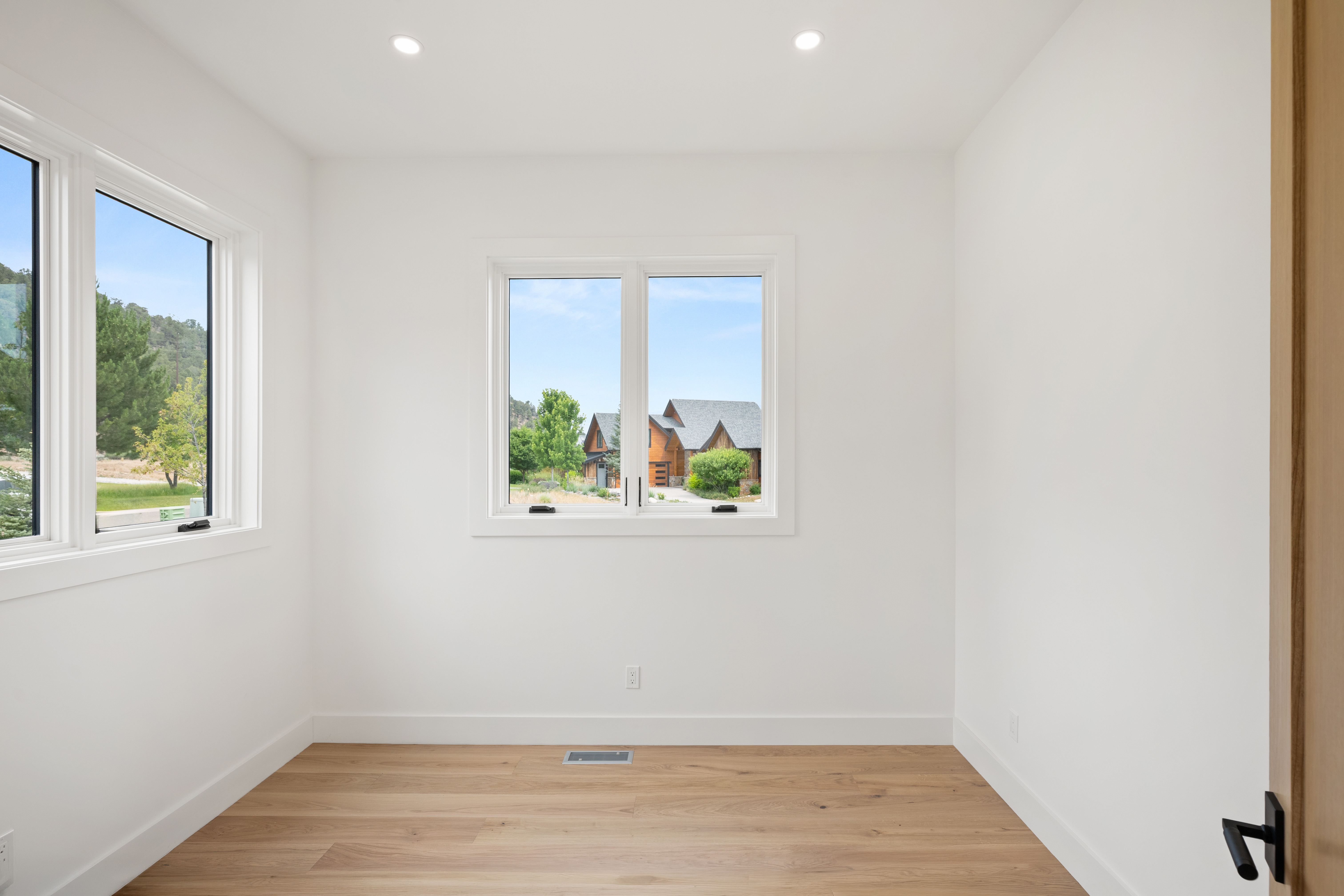 This minimalist, sunlit room features light hardwood flooring and crisp white walls, offering a versatile space ideal for a home office or study. Large windows provide natural light and a pleasant view of the surrounding residential neighborhood, creating an inviting and peaceful atmosphere. The clean lines and neutral palette make it a blank canvas ready for personalization.