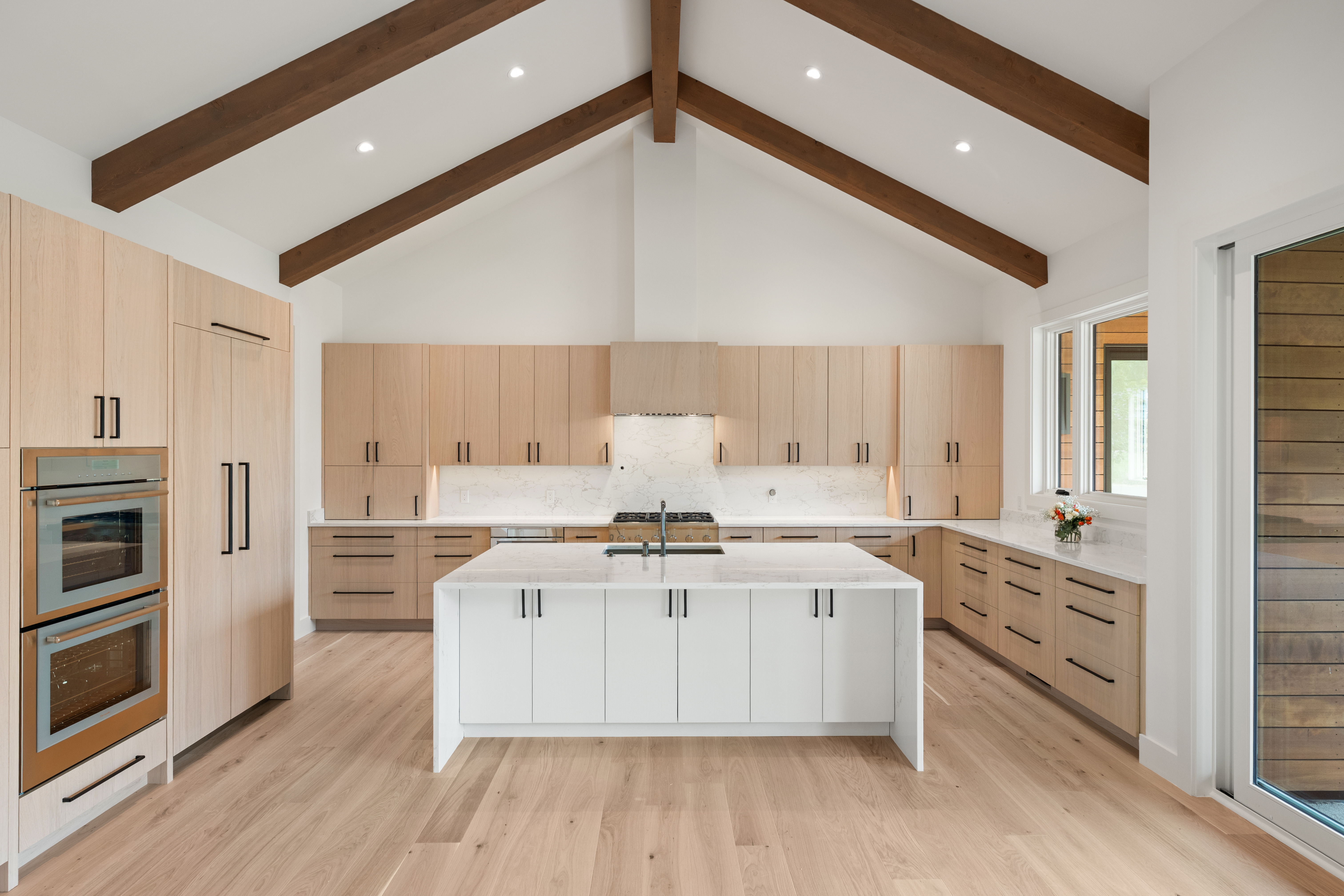 This stunning modern kitchen features a spacious open floor plan with vaulted ceilings adorned with rustic exposed wooden beams. It boasts custom light-wood cabinetry, a large white quartz-topped central island, and high-end stainless steel appliances, including a dual-oven setup. The room feels bright and airy, complemented by light hardwood flooring and large windows that provide a seamless connection to the outdoors.
