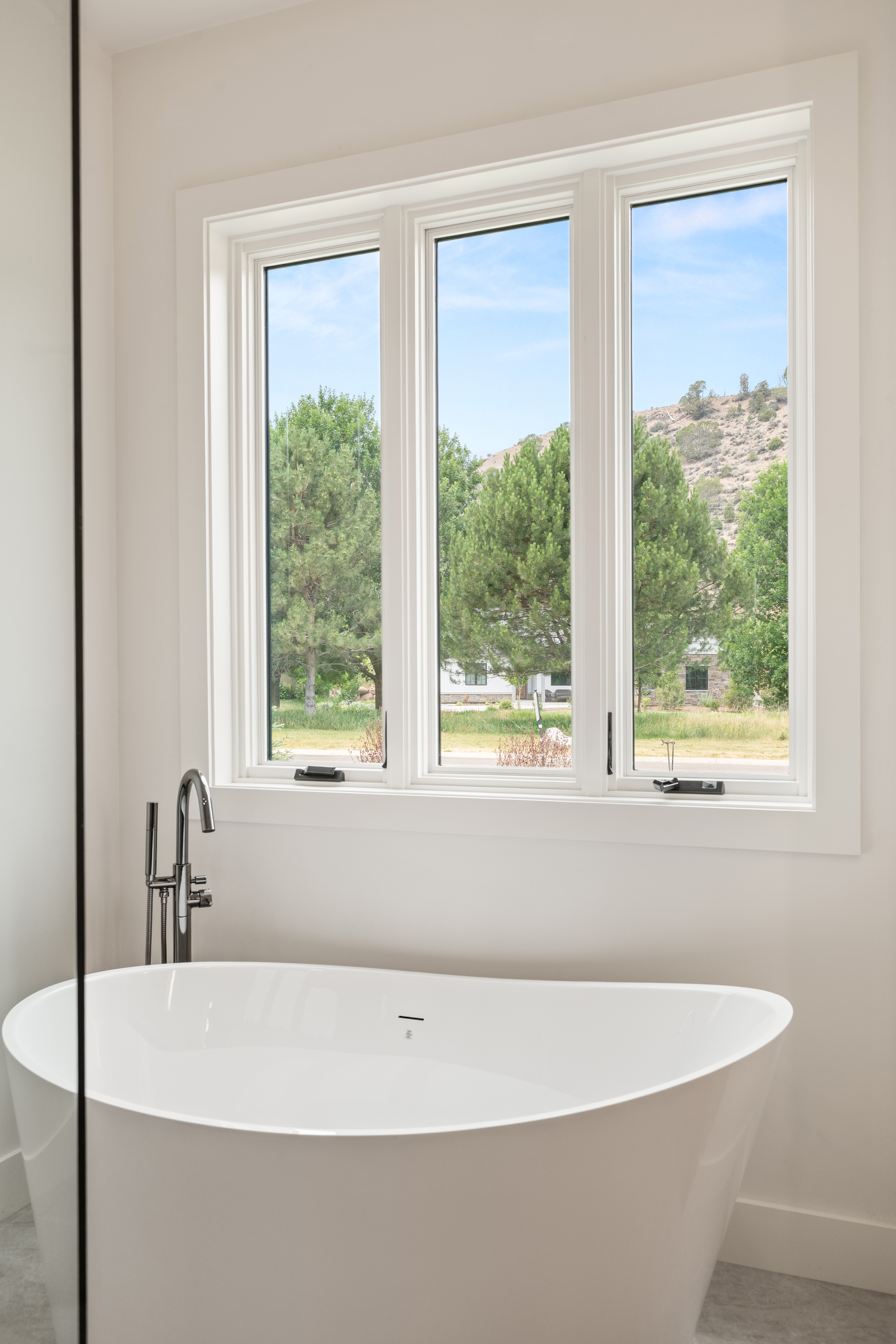This primary bathroom features a sleek, modern freestanding soaking tub positioned beneath a large three-pane casement window that provides abundant natural light and scenic views. The minimalist design is accentuated by contemporary polished chrome faucet fixtures, creating a clean and tranquil spa-like atmosphere.