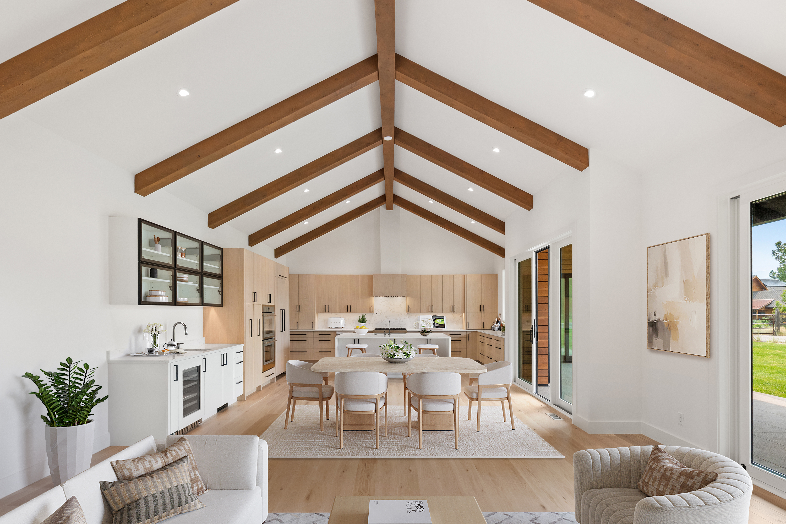 This bright, open-concept kitchen and dining area features vaulted ceilings with exposed wood beams and light wood cabinetry for a modern, organic aesthetic. The space is anchored by a large central island and a dining set, complemented by a dedicated bar area with a wine cooler. Large sliding glass doors flood the area with natural light, seamlessly connecting the interior to the outdoor surroundings.