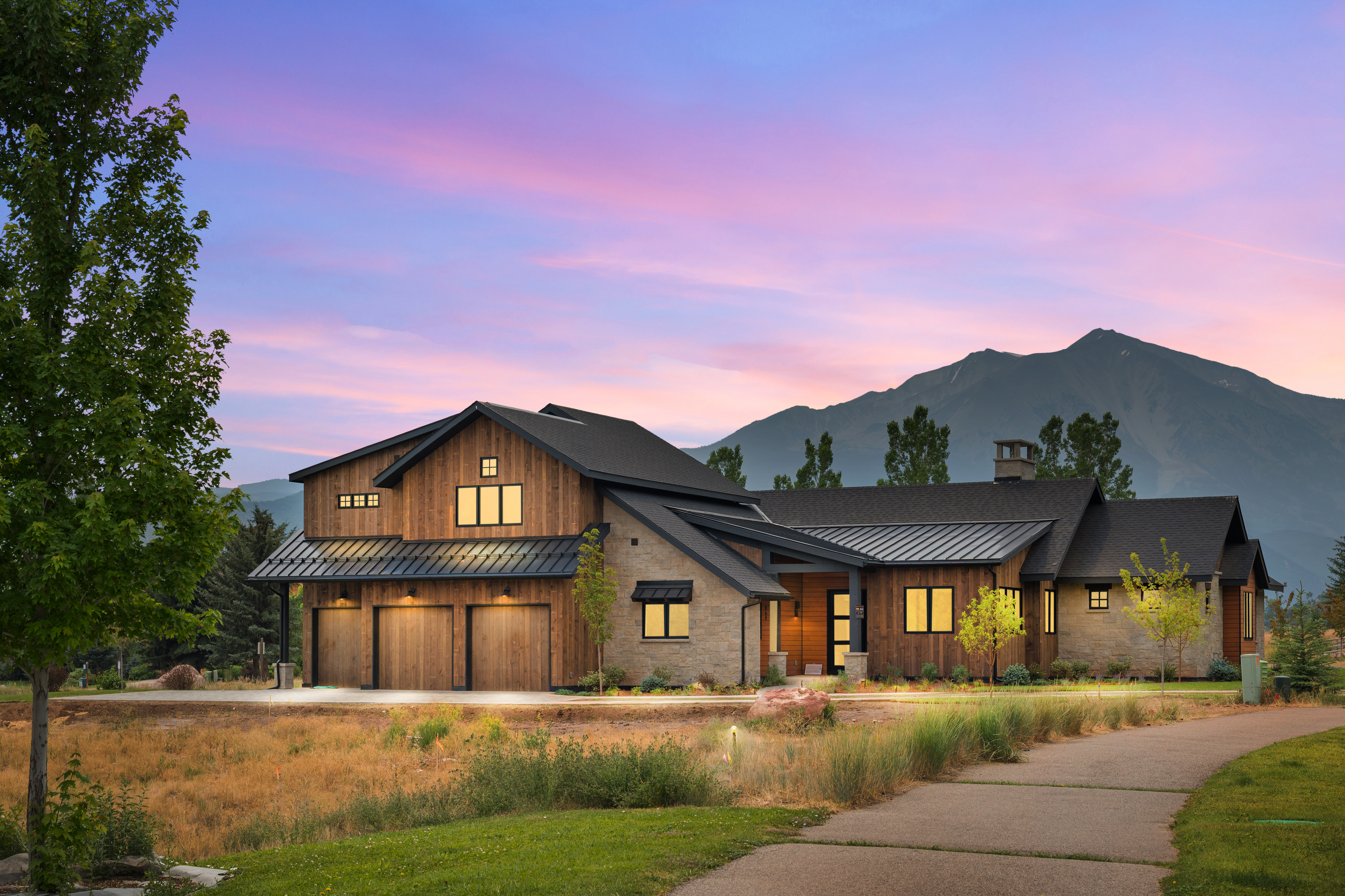 This stunning contemporary mountain home features a sophisticated blend of natural wood siding and textured stone masonry, accented by dark metal roofing and window frames. The property includes a spacious three-car garage and a welcoming entryway, all set against a breathtaking backdrop of mountain peaks at sunset. Its architectural design perfectly balances rugged rustic charm with modern luxury, making it an impressive display of high-end mountain living.