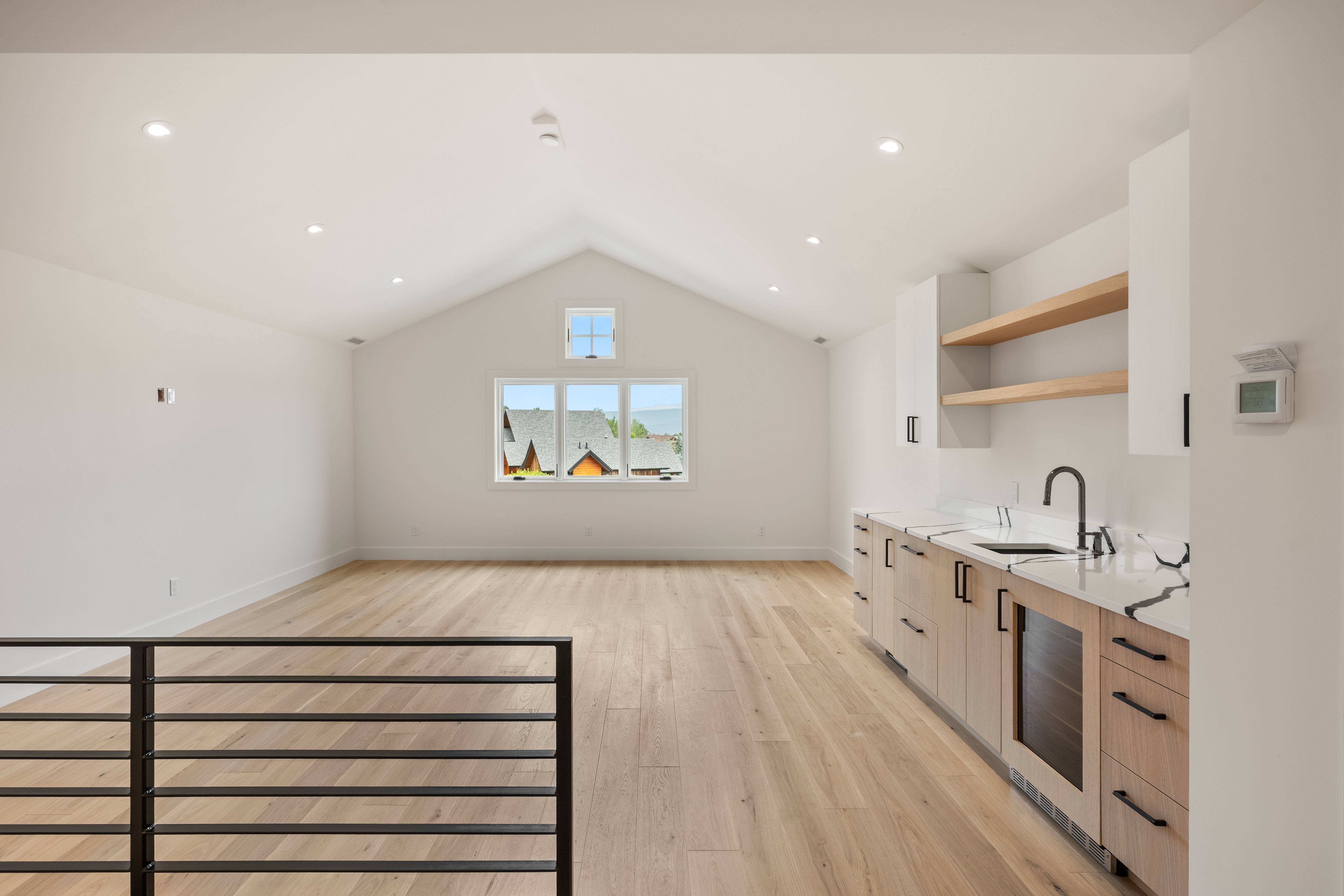 This bright, contemporary open-concept space features a kitchenette with light wood cabinetry, white marble-style countertops, and floating shelves. The room is highlighted by a vaulted ceiling with recessed lighting and large windows that provide abundant natural light. The aesthetic is clean and modern, offering a versatile space that could serve as a living area or lounge.