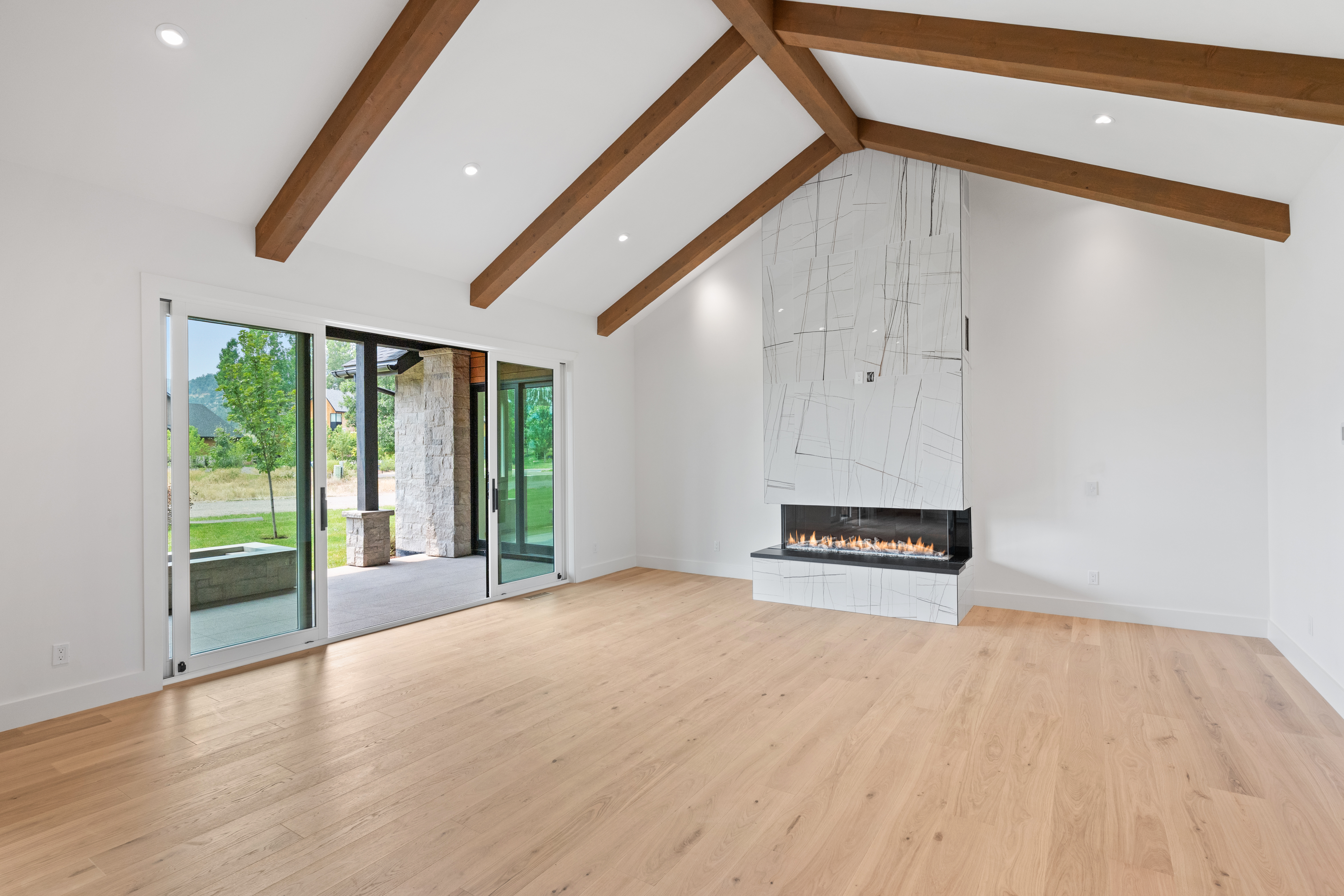 This spacious, modern living area features high vaulted ceilings with exposed wood beams and light hardwood flooring throughout. A striking floor-to-ceiling fireplace with marble tile cladding serves as the focal point of the room. Large glass sliding doors lead out to a patio, flooding the space with natural light and providing a seamless transition to the outdoors.