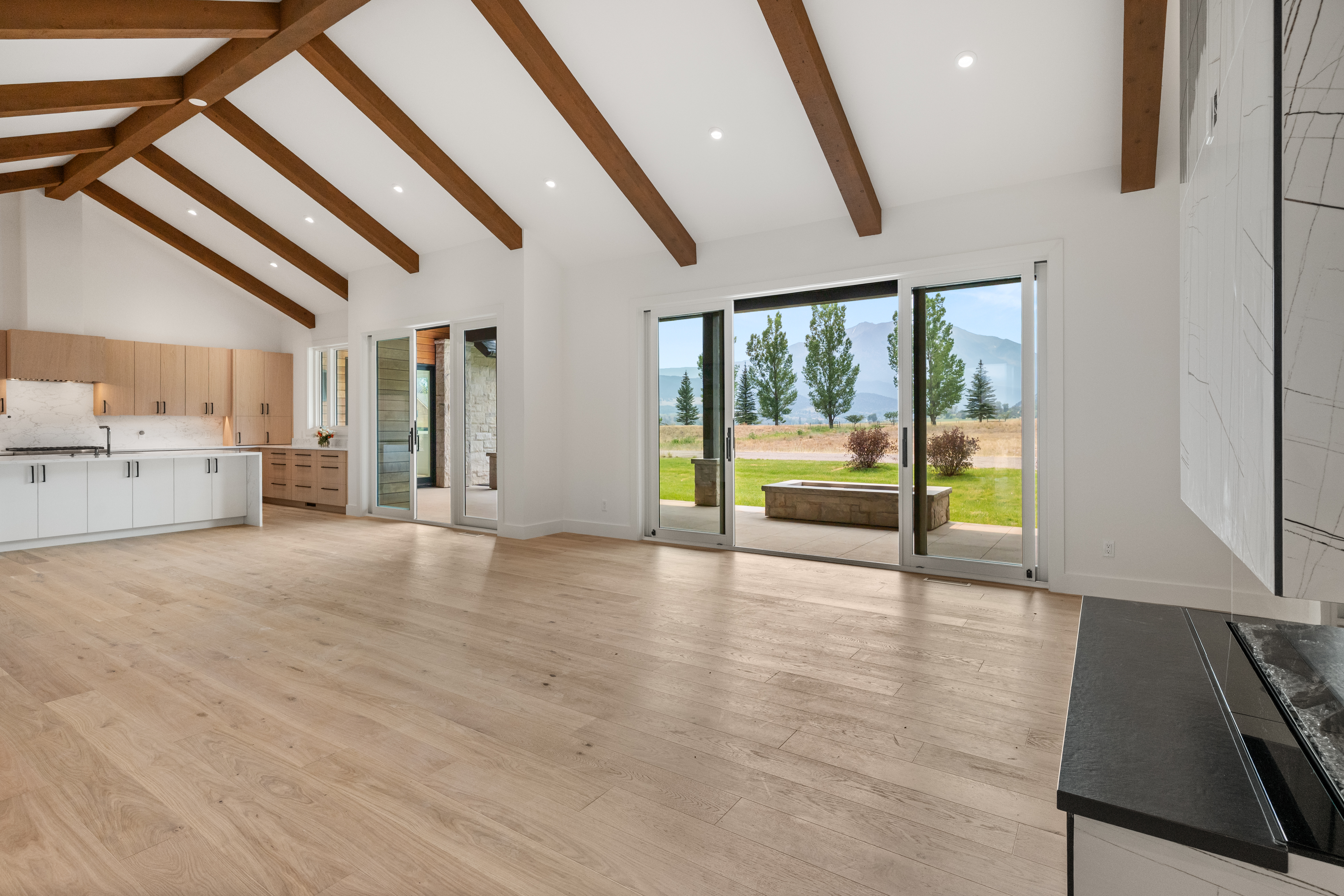 This bright, open-concept space features a modern kitchen with light wood upper cabinetry and white lower cabinets, seamlessly flowing into a spacious living area. High vaulted ceilings with exposed wood beams add architectural character, while large sliding glass doors provide expansive views of the outdoor landscape. The interior aesthetic is minimalist and airy, emphasizing clean lines and high-quality finishes.