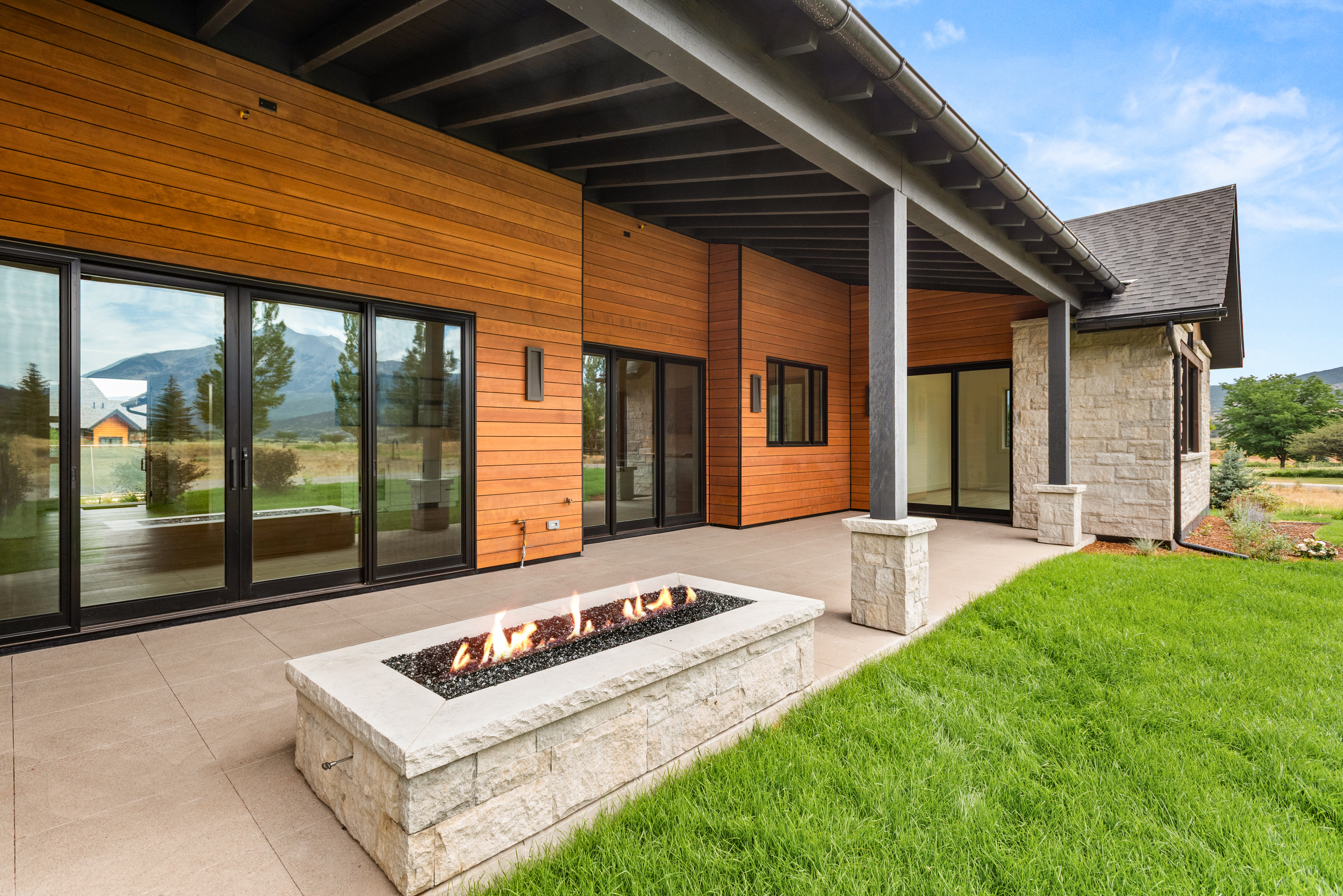 This modern covered patio space features smooth tile flooring and a built-in stone fire pit, creating an ideal setting for outdoor entertaining. The exterior combines sleek horizontal wood paneling with rugged stone accents, reflecting a sophisticated, contemporary rustic design. Large sliding glass doors provide seamless indoor-outdoor flow while offering scenic views of the surrounding landscape.