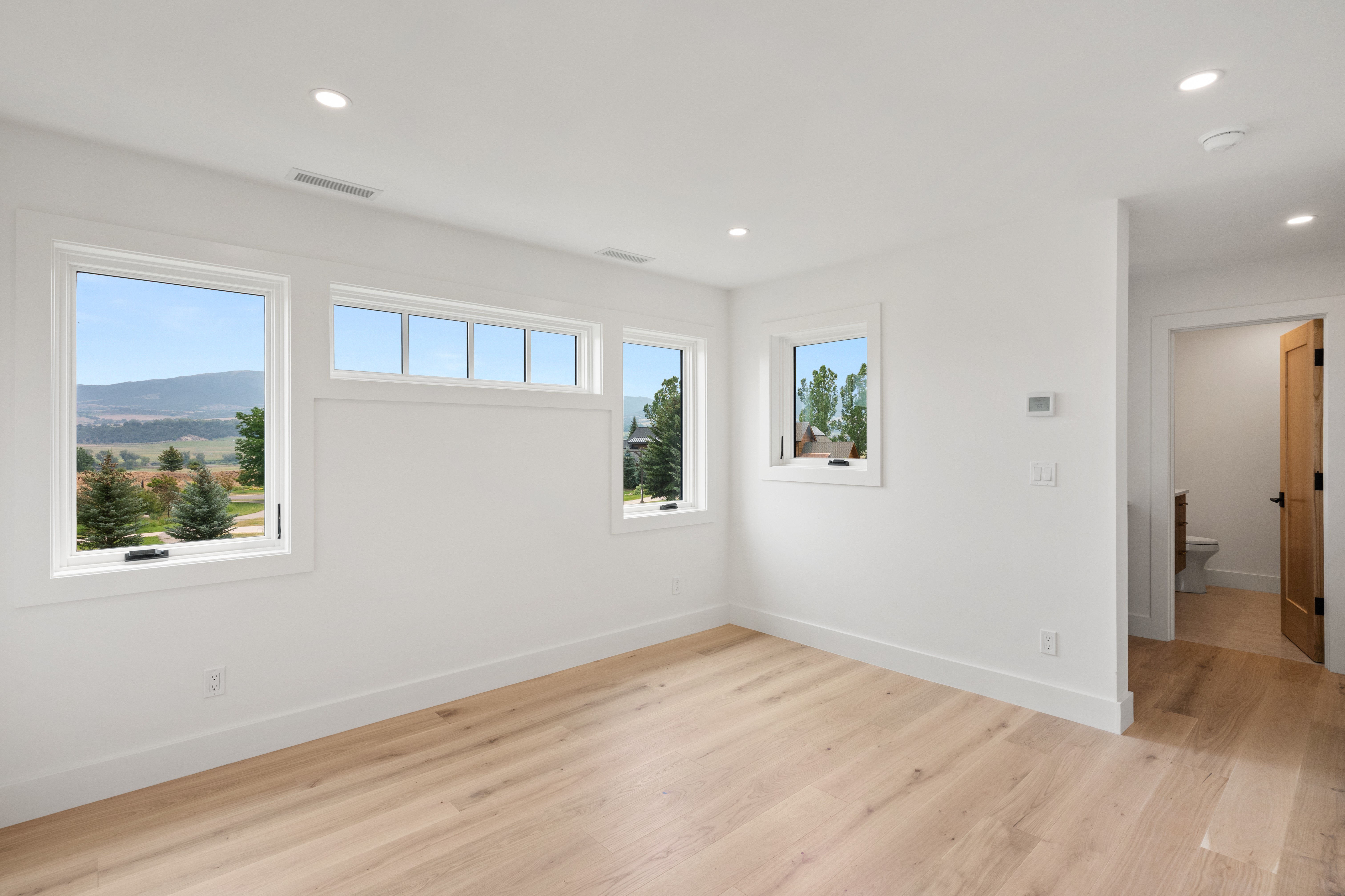 A bright and minimalist bedroom featuring light wood flooring and crisp white walls. The space offers abundant natural light through multiple windows, including a large picture window with scenic mountain and tree views. A doorway leads to an adjoining bathroom, highlighting a functional and clean layout.