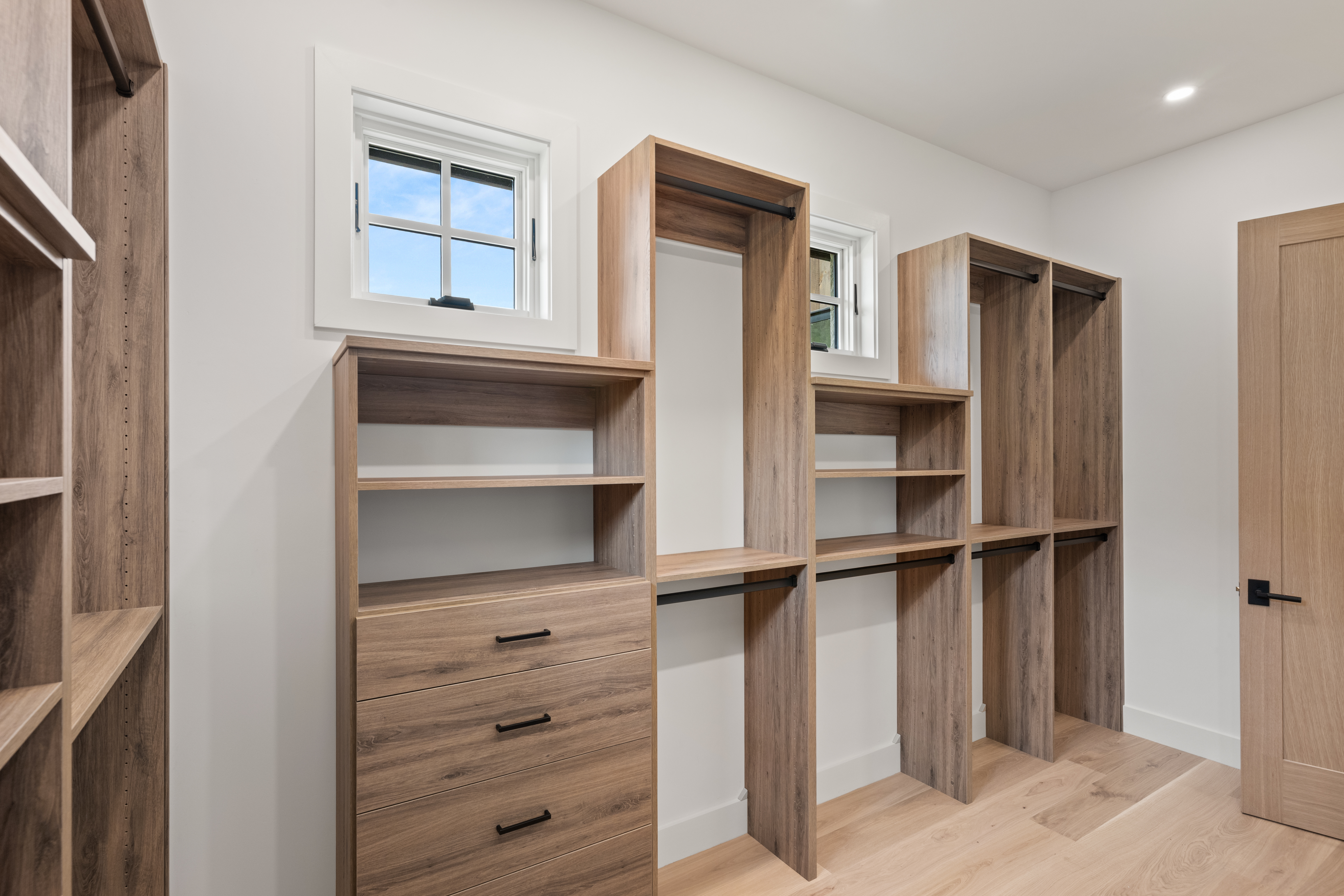 This spacious and well-organized walk-in closet features custom wood-toned shelving and built-in drawers, offering ample storage space for clothing and accessories. Two windows allow for natural light to fill the room, highlighting the clean, modern design and light-colored walls. The layout includes multiple hanging areas and open shelving, making it a functional and stylish addition to the home.