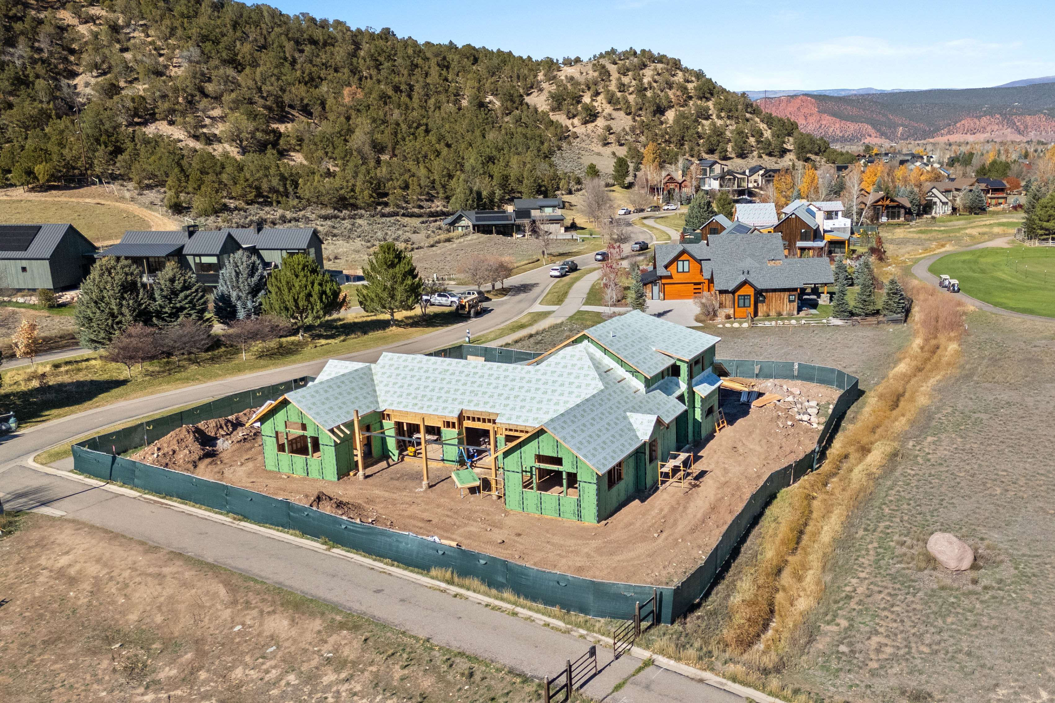 This aerial view showcases a residential property under construction in a serene, scenic community. The new build, framed and sheathed with green weather-resistant panels, features a sprawling floor plan set against a backdrop of rolling hills and established modern homes. The site is securely enclosed by fencing, emphasizing the ongoing development within this desirable neighborhood setting.