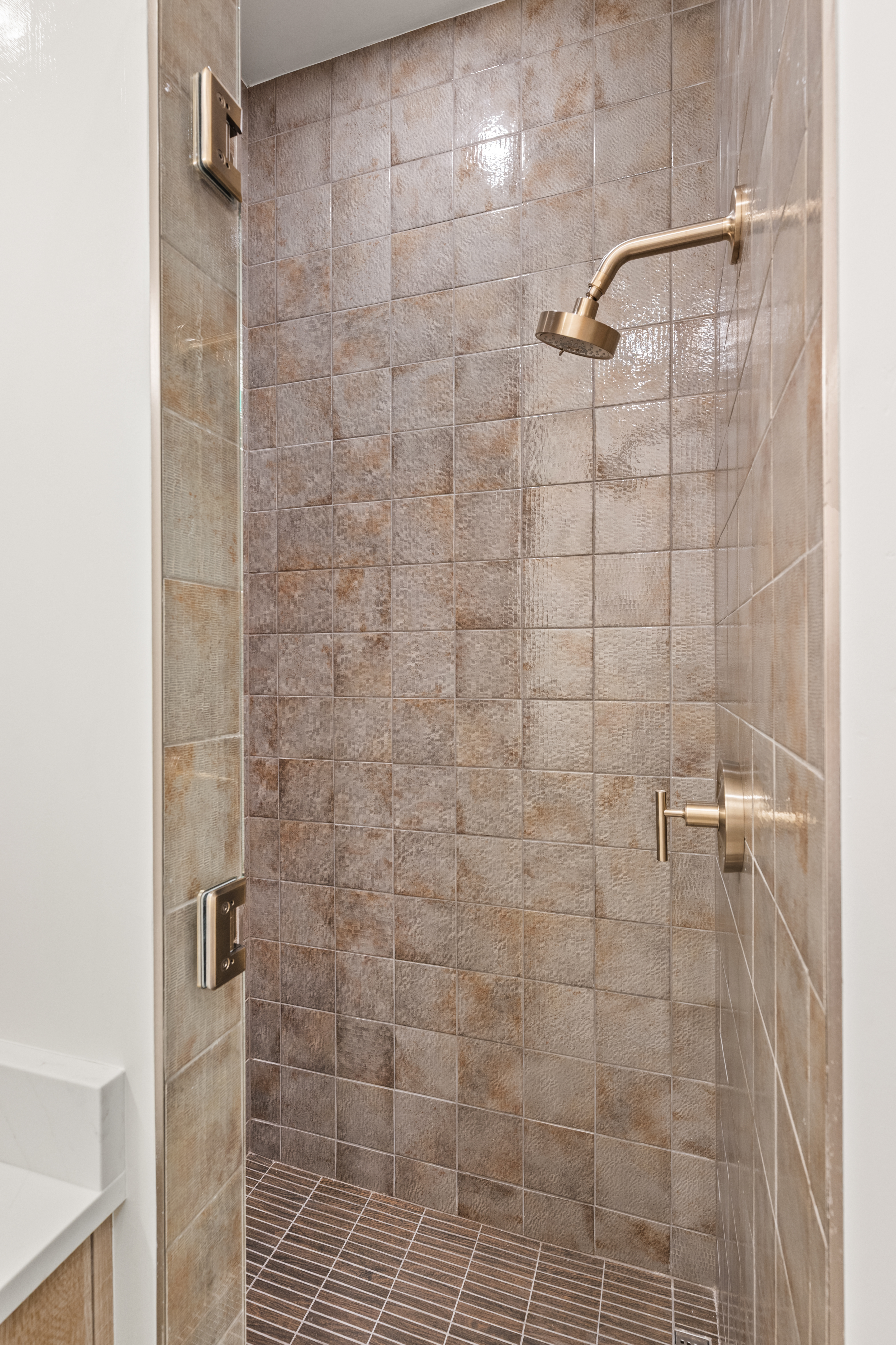 This walk-in shower features textured, earthy-toned square tiles that create a warm and rustic aesthetic. It is equipped with contemporary brushed gold fixtures, including a rainfall showerhead and a sleek handle control, complemented by a glass panel door. The floor uses contrasting linear mosaic tiles, adding a sophisticated, modern touch to the space.