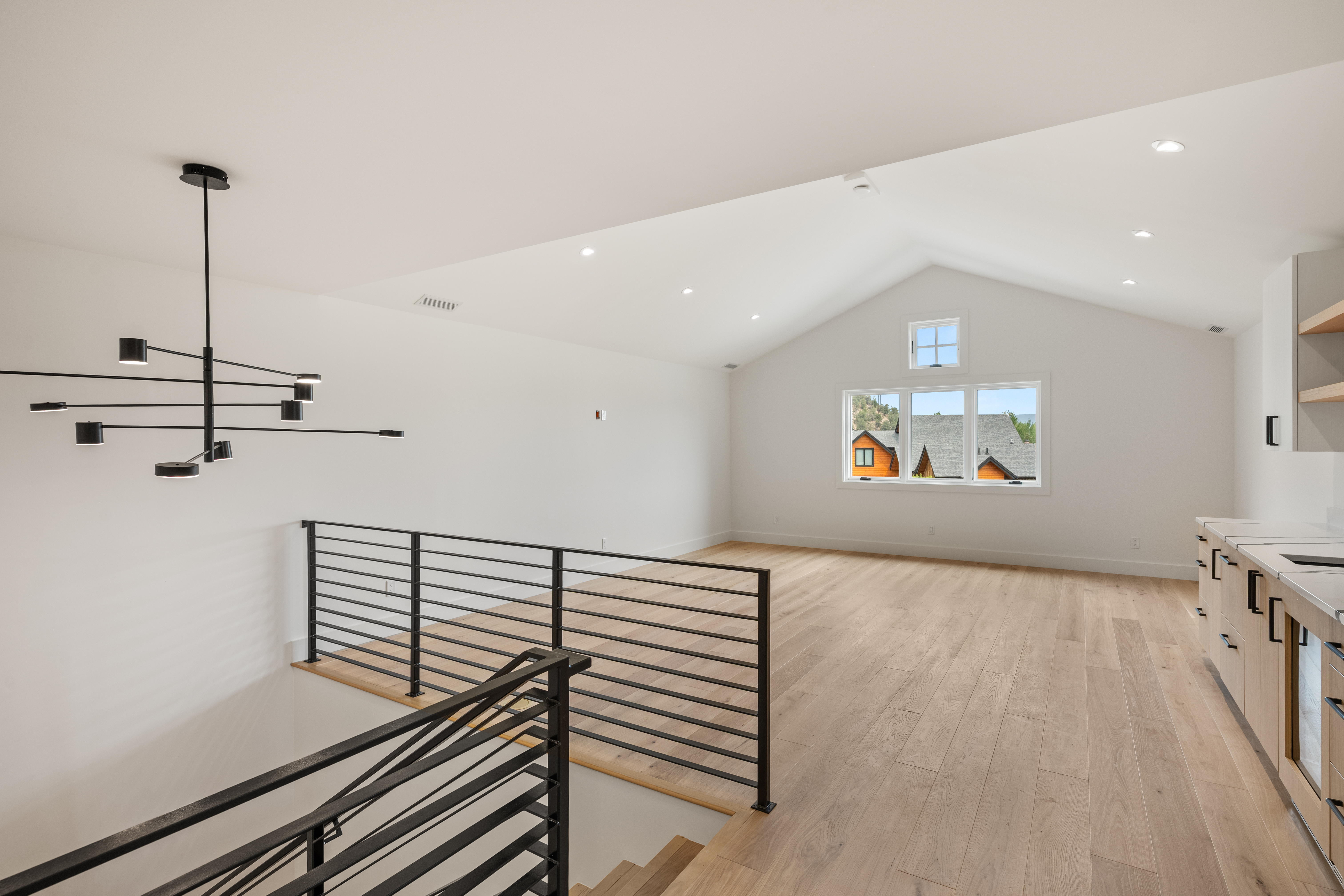 This spacious, open-concept loft area features high vaulted ceilings and light wood flooring, creating an airy and modern atmosphere. A striking geometric chandelier hangs over the landing, which is protected by a sleek black metal railing that overlooks the stairs. To the right, a kitchenette with light wood cabinetry and dark hardware adds functionality to this versatile living space.