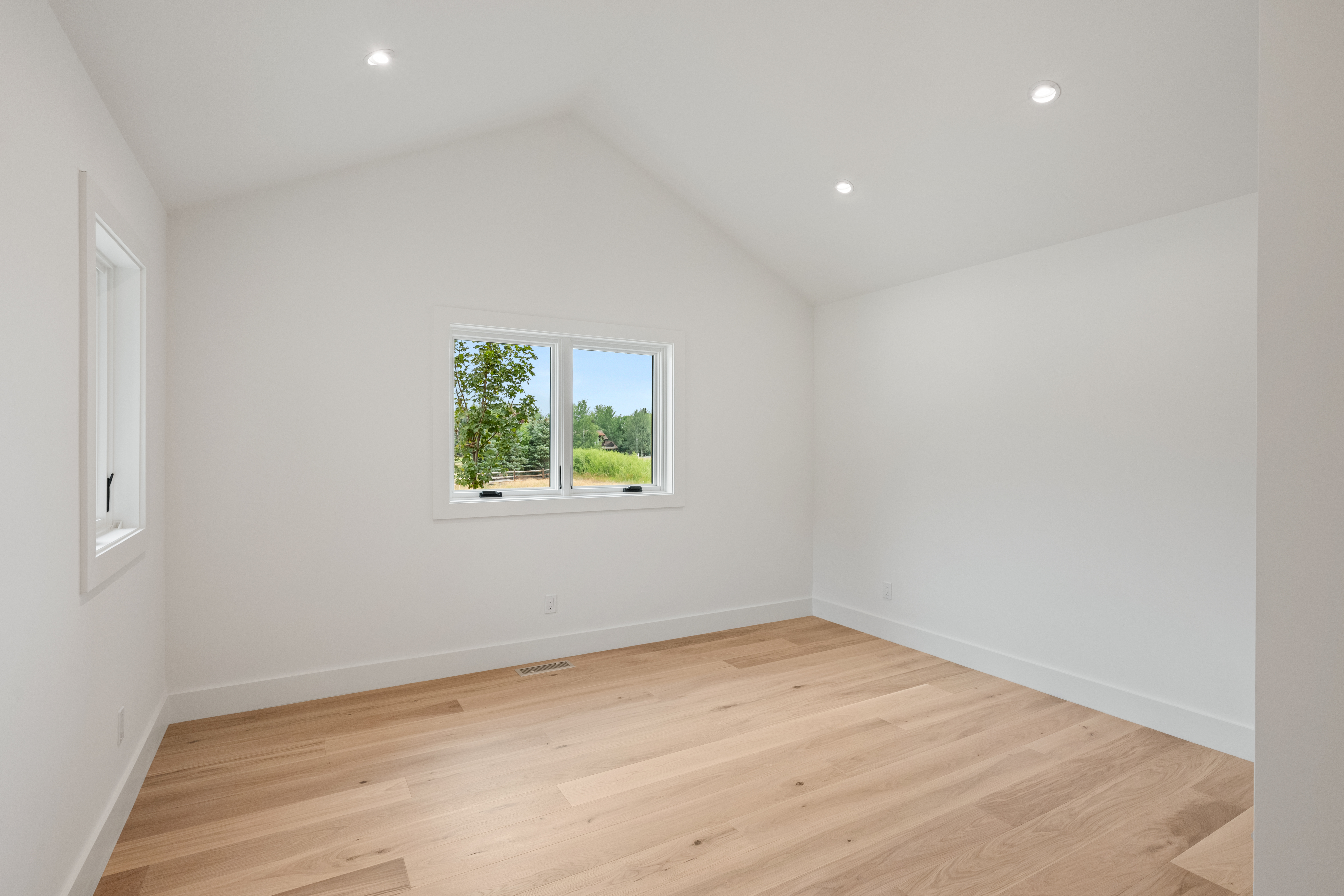 This is a clean, bright, and empty bedroom featuring high vaulted ceilings and light-toned hardwood flooring. The room is neutrally painted with white walls and baseboards, creating a versatile blank canvas for any design style. A double-pane window provides natural light and an pleasant view of the surrounding greenery.