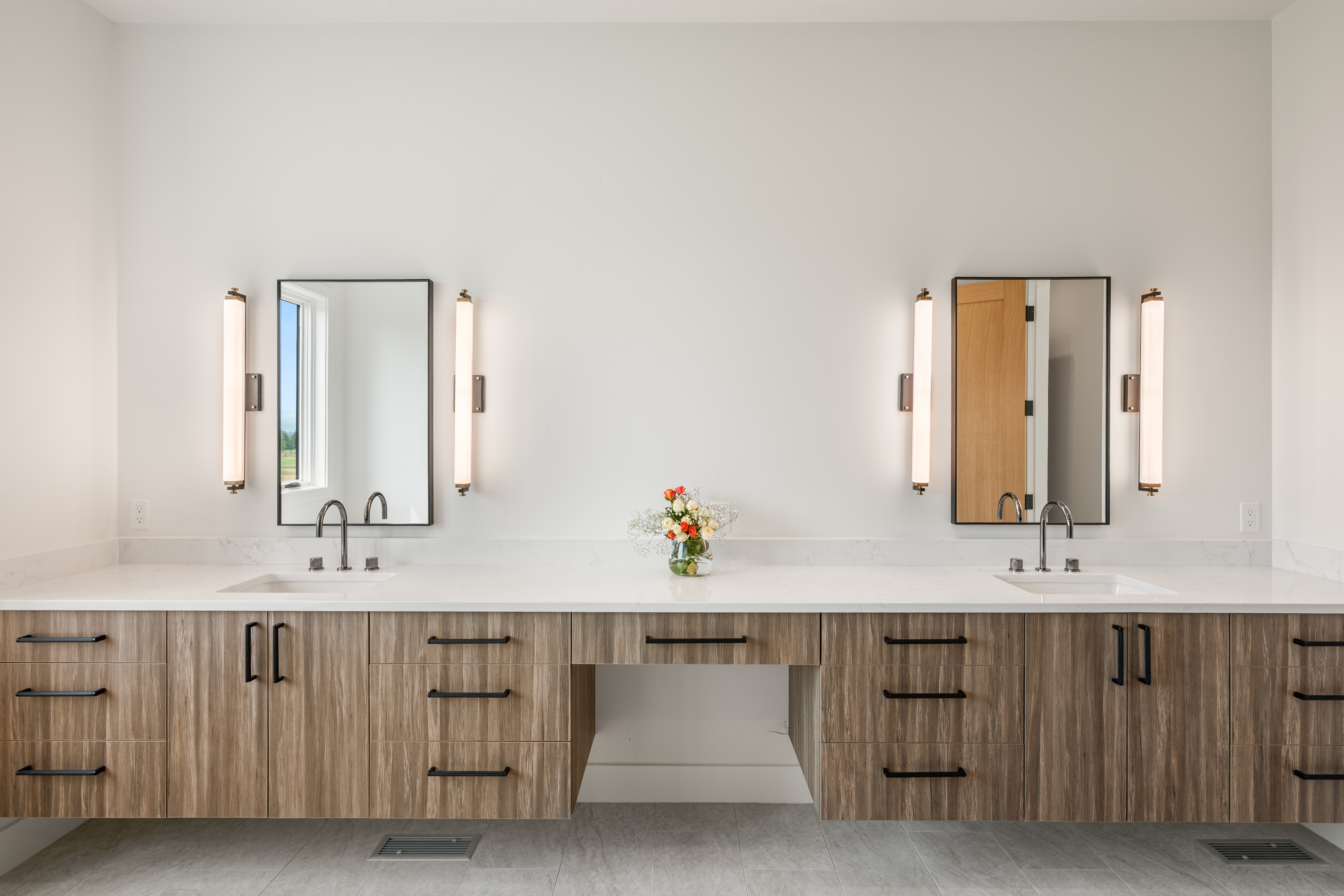 This modern primary bathroom features a double-vanity with rich wood-toned cabinetry, a clean white quartz countertop, and minimalist black hardware. The space is highlighted by two rectangular mirrors flanked by sleek, vertical wall sconces and a central vanity knee space with a fresh floral arrangement. The overall aesthetic is contemporary and sophisticated, offering a bright, airy, and functional design.