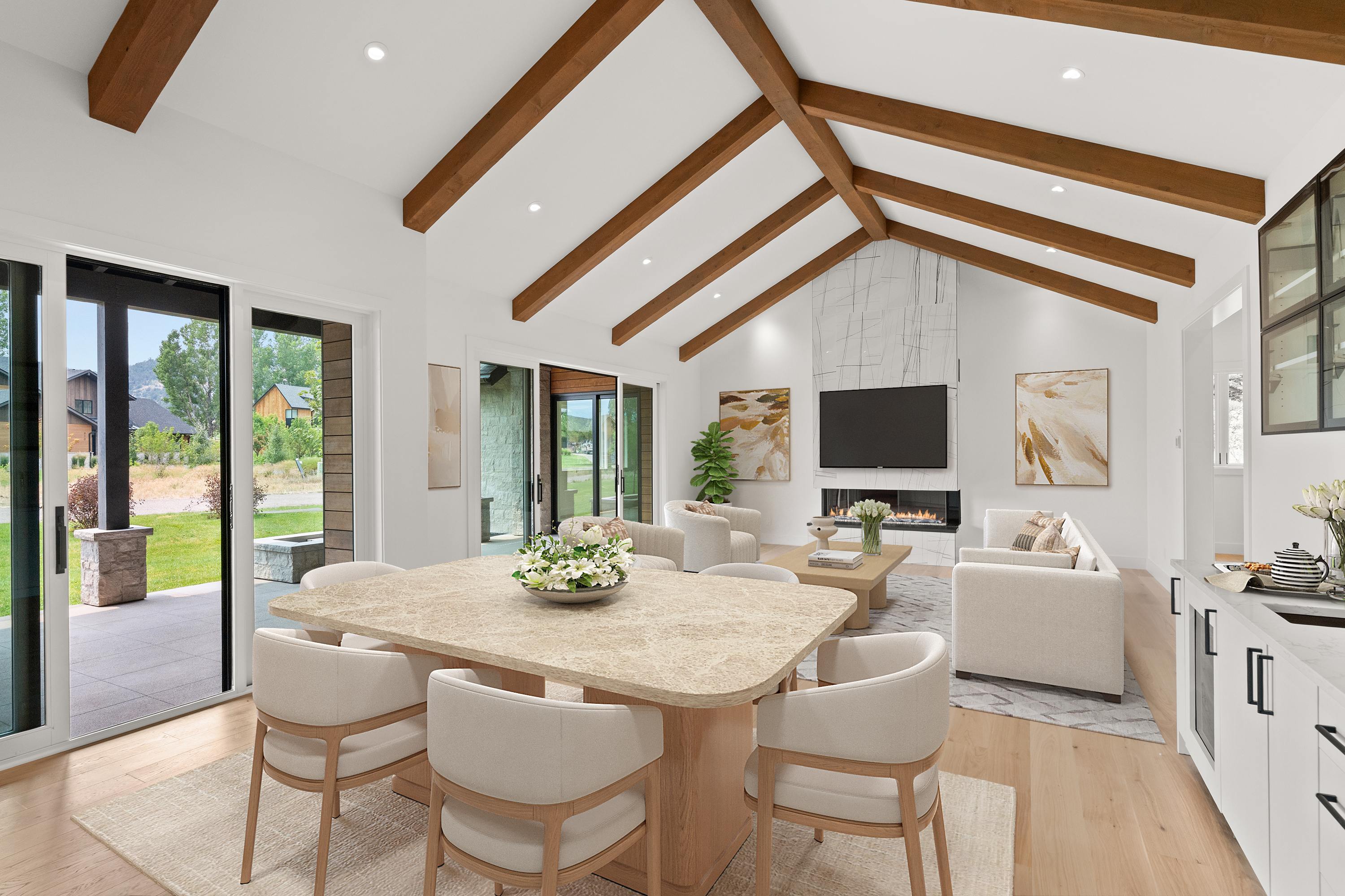 This bright, open-concept space features a contemporary dining area that flows seamlessly into a spacious living room with a vaulted ceiling and striking exposed wood beams. The room is highlighted by a large stone fireplace, a neutral color palette, and expansive glass sliding doors that provide abundant natural light and outdoor views. The high-end finishes and clean architectural lines create a sophisticated yet inviting atmosphere.