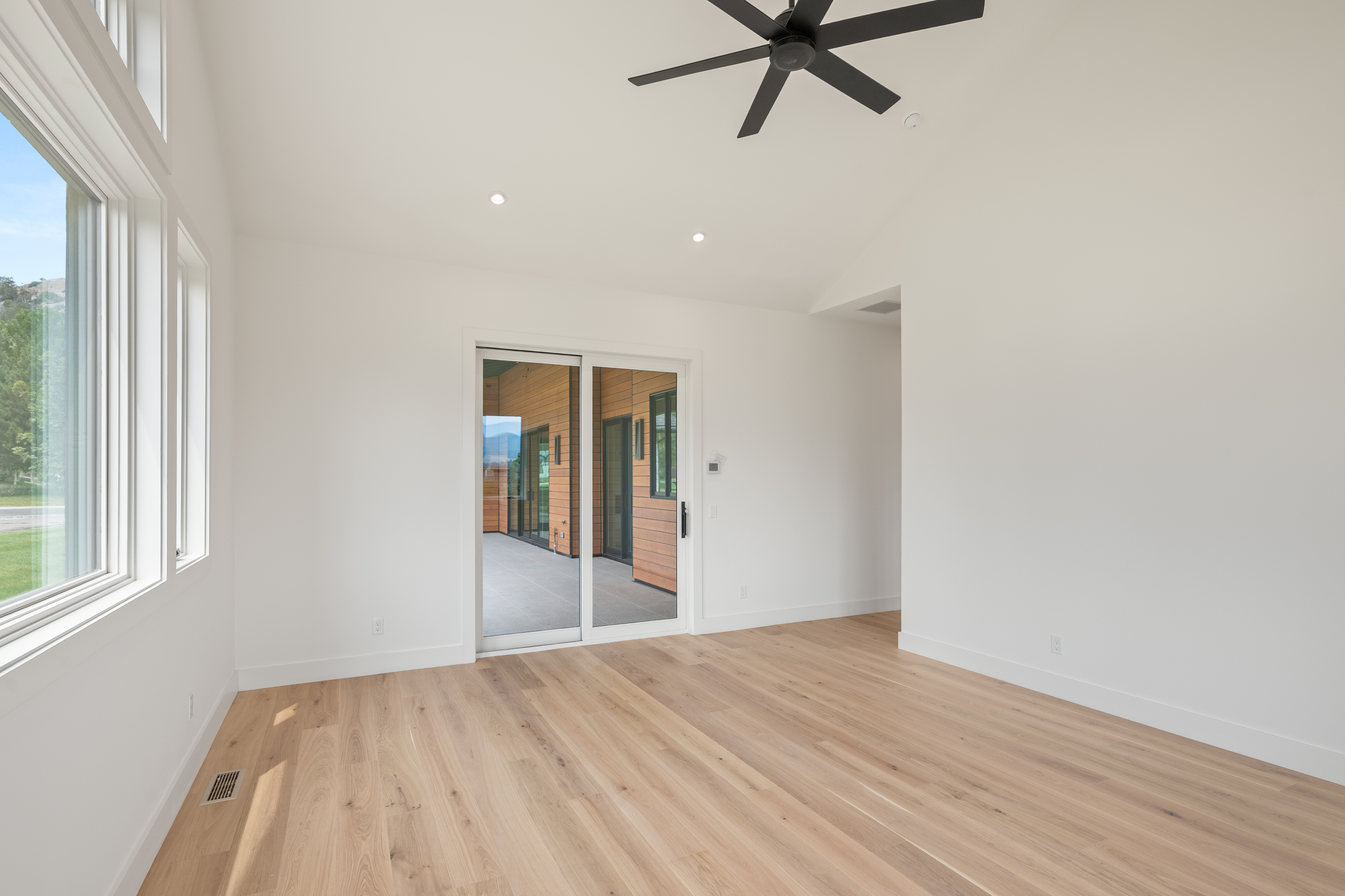 This is a bright and minimalist interior space, likely a bedroom, featuring light-toned hardwood flooring and crisp white walls. The room is accented by a modern black ceiling fan and large windows that provide abundant natural light, along with a sliding glass door that opens to a covered patio. The clean, contemporary aesthetic creates an airy and inviting atmosphere.
