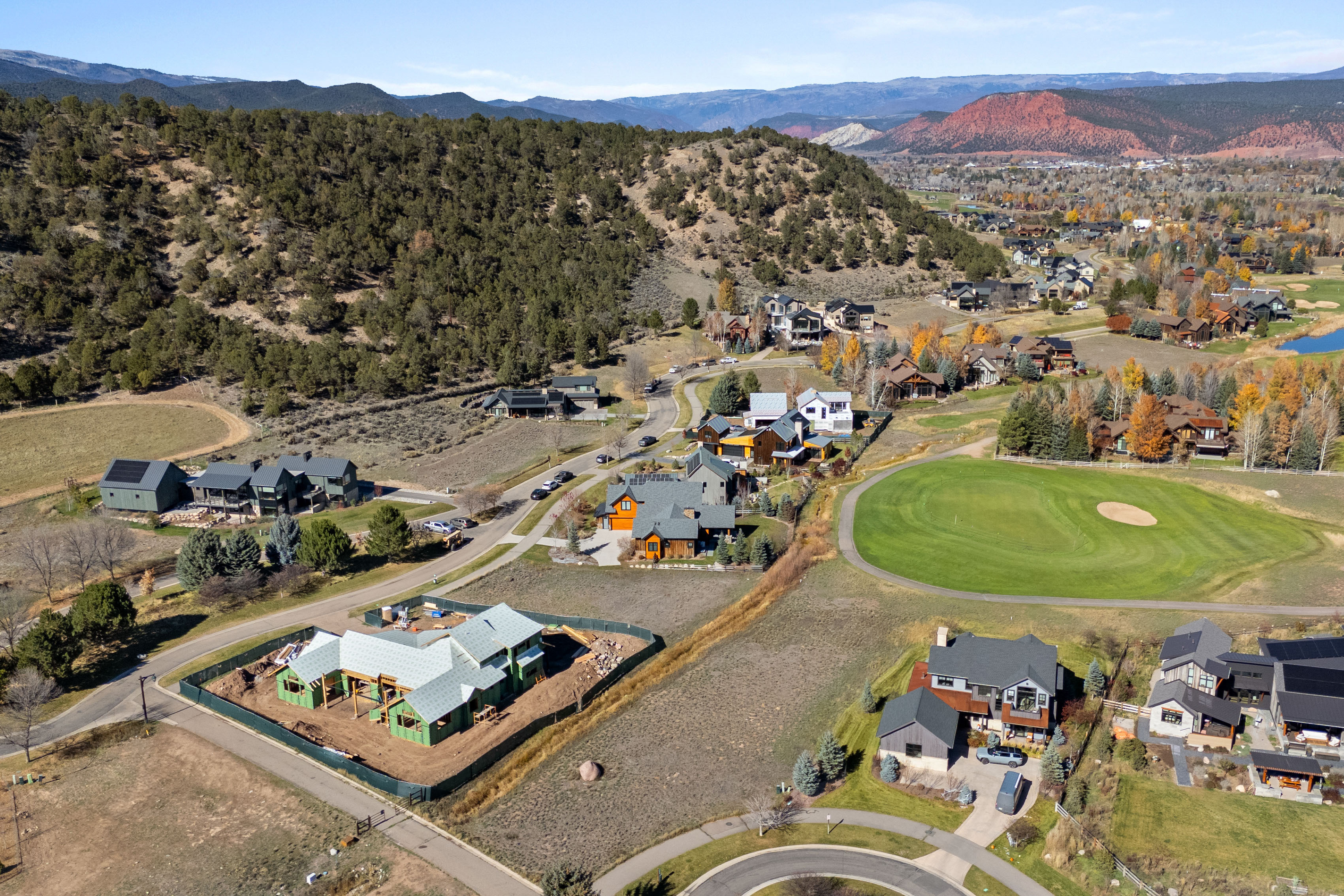 This aerial photograph captures a scenic, upscale residential development nestled against a backdrop of rolling hills and majestic mountains. The neighborhood features a mix of modern and traditional architectural styles, including a prominent construction site in the foreground and a well-manicured golf course green. The community exhibits a cohesive, high-end feel with spacious lots and natural, landscaped surroundings.