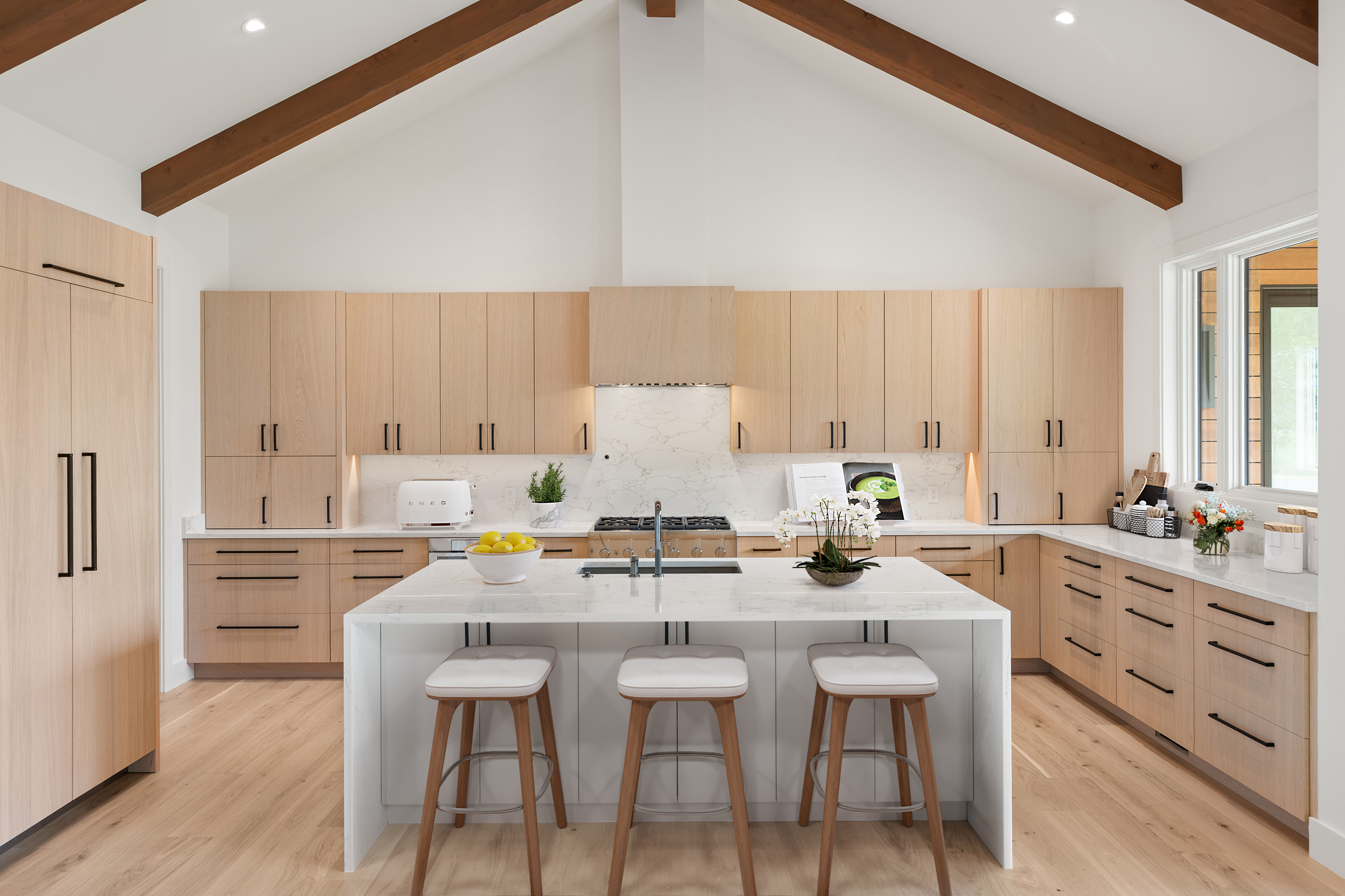 This modern kitchen features warm wood cabinetry, a waterfall-edge marble island, and high-end stainless steel appliances. The high vaulted ceiling with exposed wooden beams creates an airy, spacious feel, while the neutral color palette provides a sophisticated, clean aesthetic. The well-appointed layout includes ample storage and integrated lighting, making it a functional and inviting centerpiece for the home.