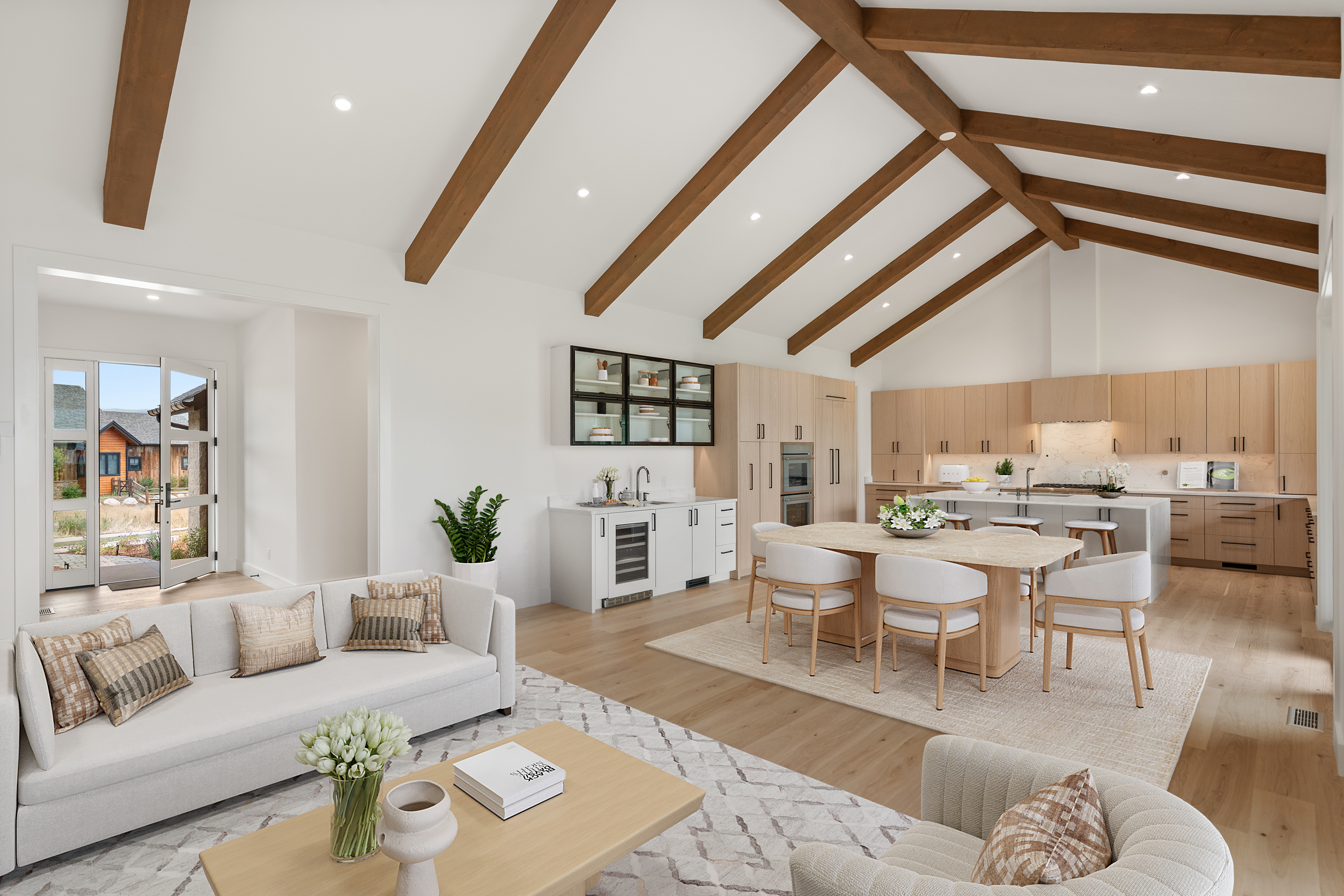 This spacious, open-concept living area features a dramatic vaulted ceiling with exposed wooden beams and light-toned hardwood flooring throughout. The space seamlessly integrates a sophisticated seating area with a formal dining space and a gourmet kitchen featuring light wood cabinetry and a large stone-topped island. Large glass doors open to an outdoor view, bathing the bright, neutral-toned interior in abundant natural light, creating an airy and luxurious atmosphere.