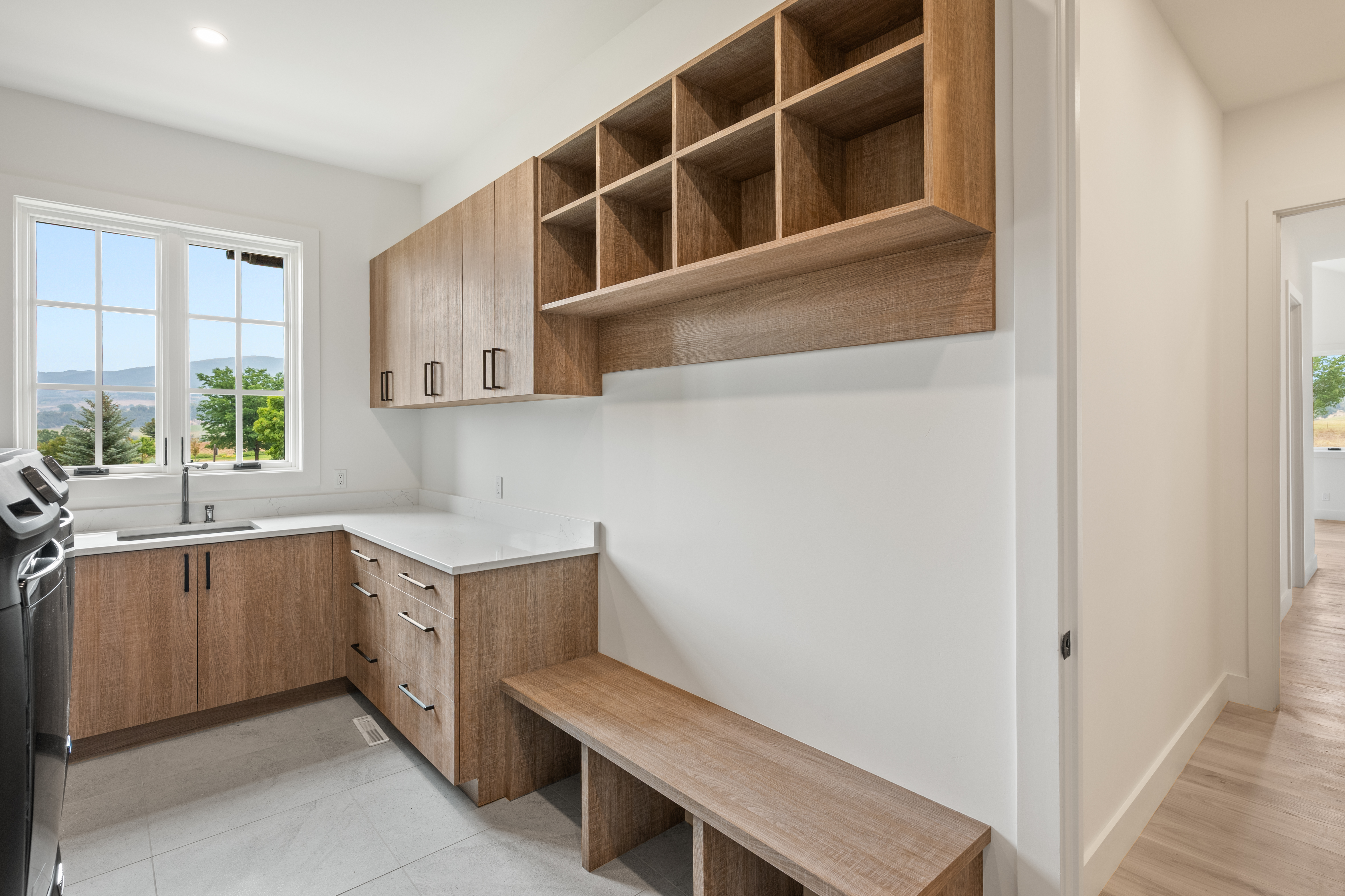 This modern laundry room features sleek wood-toned cabinetry with a clean white countertop and an integrated sink. The space is efficiently designed with built-in storage cubbies above a matching bench seat, providing both functionality and organization. With its neutral color palette and bright lighting, the room offers a clean and contemporary utility space.