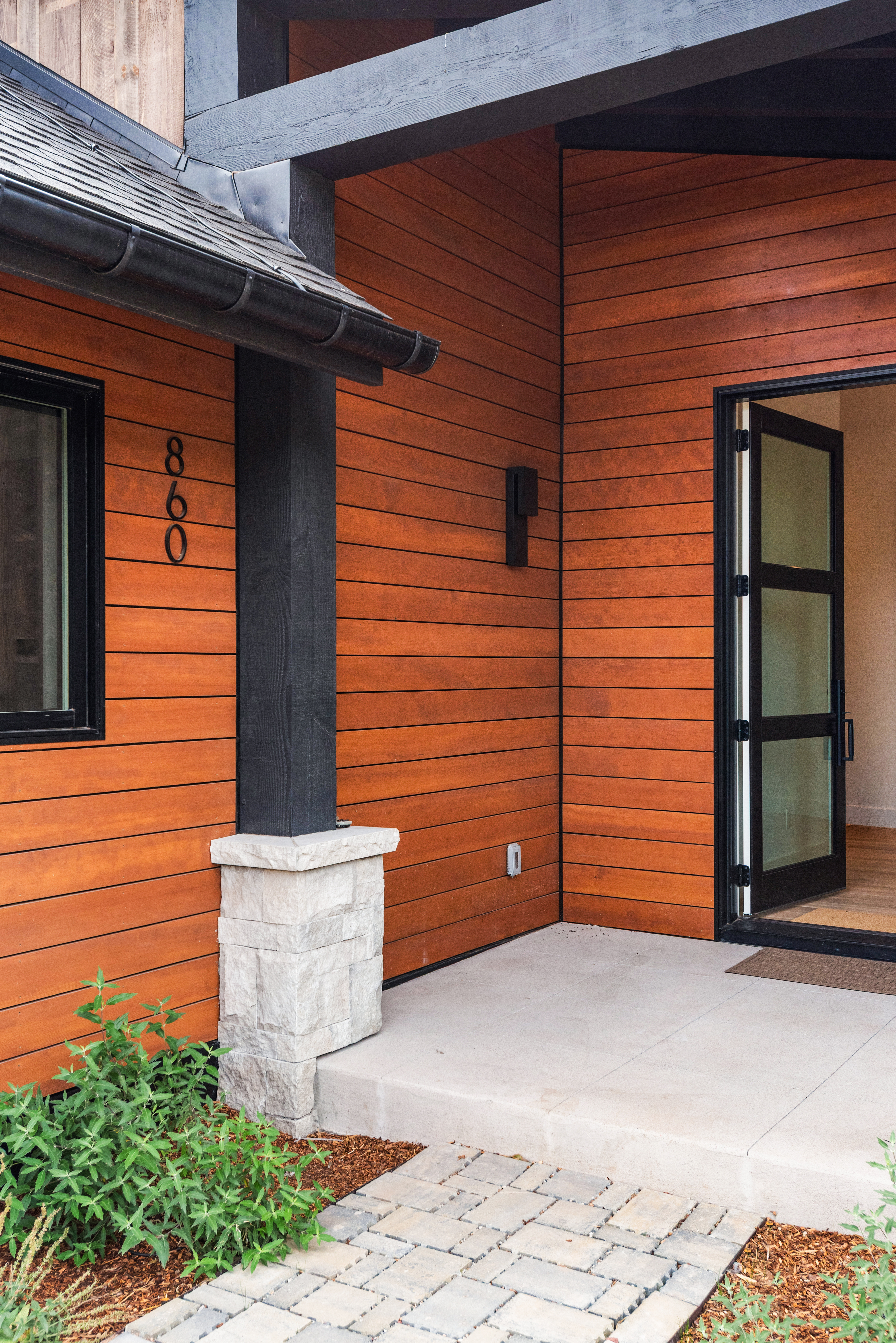 This modern entryway features warm horizontal wood siding contrasted by sharp black trim and a structural post with a stone base. The house number '860' is displayed vertically beside a large window, while a contemporary glass-paneled door opens into the home. A clean concrete porch step and stone paver walkway create a welcoming and well-manicured approach.