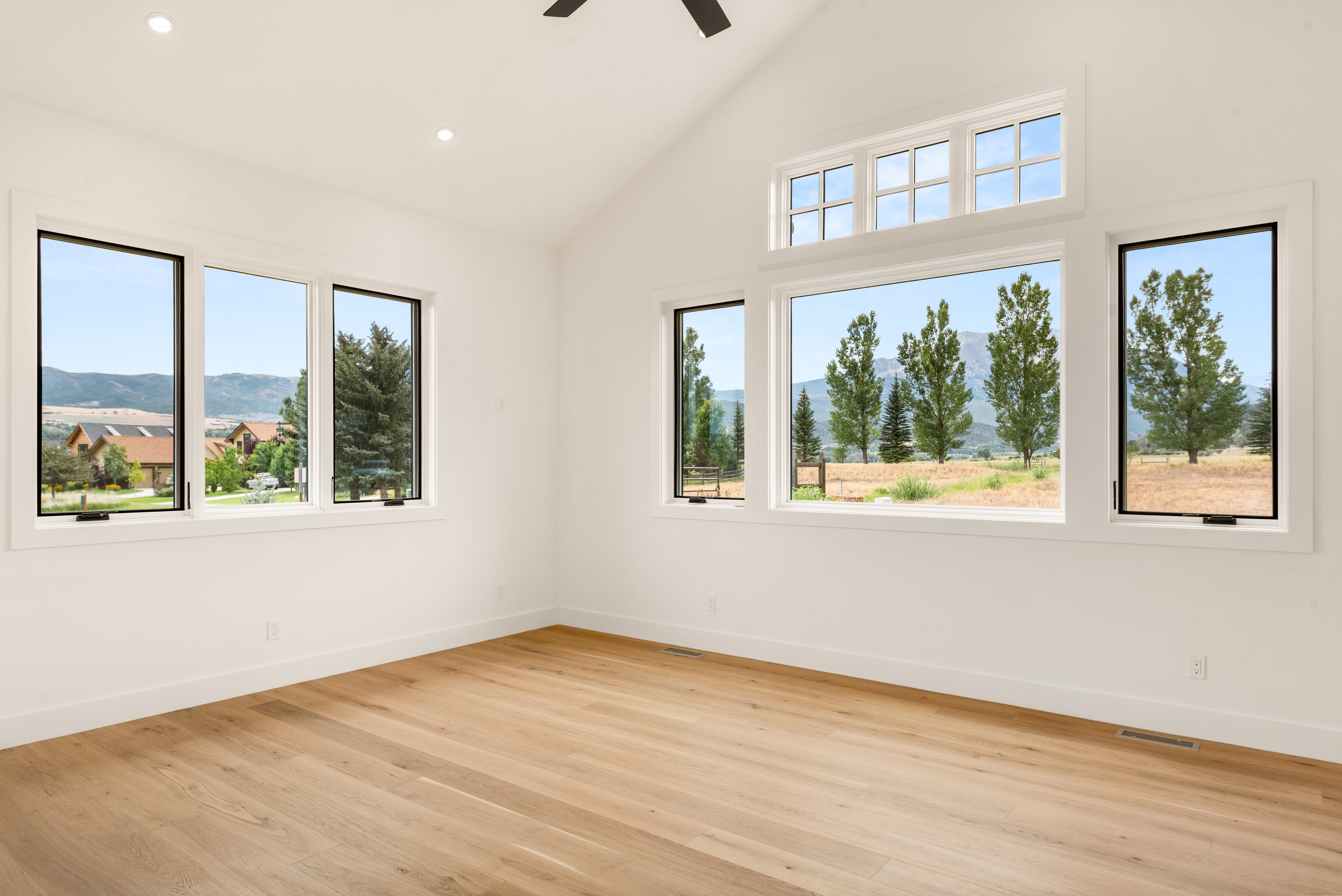 This spacious, modern living room is characterized by its bright, vaulted ceilings and light-toned hardwood flooring. Multiple large windows allow for an abundance of natural light and offer scenic views of the surrounding rural landscape. The neutral white color palette provides a clean, airy, and inviting canvas for potential homeowners.