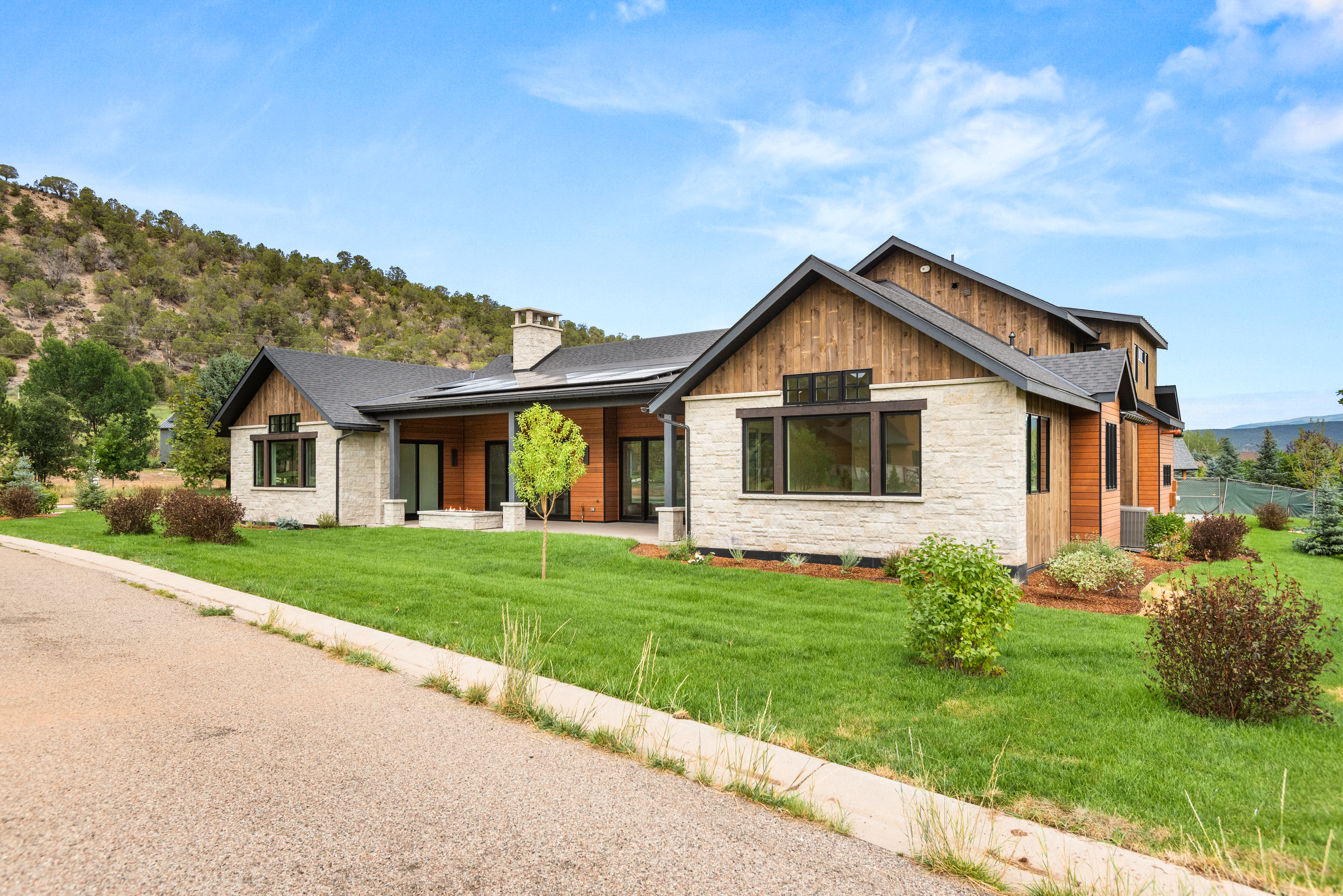 This modern farmhouse-style residence features a stunning combination of light-colored stone veneer and warm wood siding, accented by dark window frames and a clean, gabled roofline. The home is set against a scenic, hilly landscape with a well-manicured lawn and a paved driveway leading up to the property. Its inviting exterior design balances rustic charm with contemporary architectural elements, creating an elegant and welcoming curb appeal.