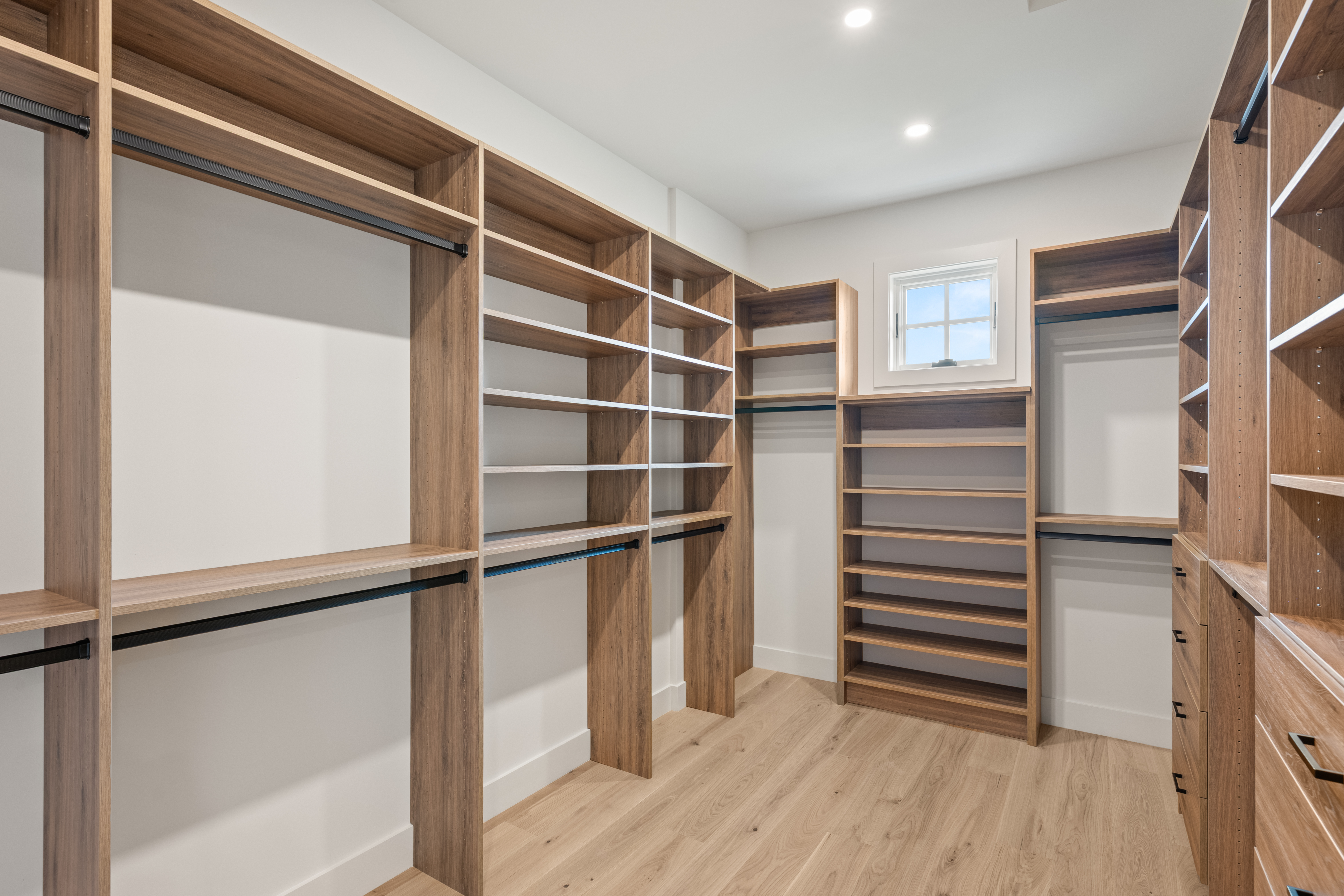 This spacious and organized walk-in closet features custom wood-toned shelving and hanging rods, providing ample storage space. The room is accented with light-colored flooring and a small, centrally positioned window that brings in natural light. Its clean, modern aesthetic and functional layout make it a highly desirable feature for a primary suite.