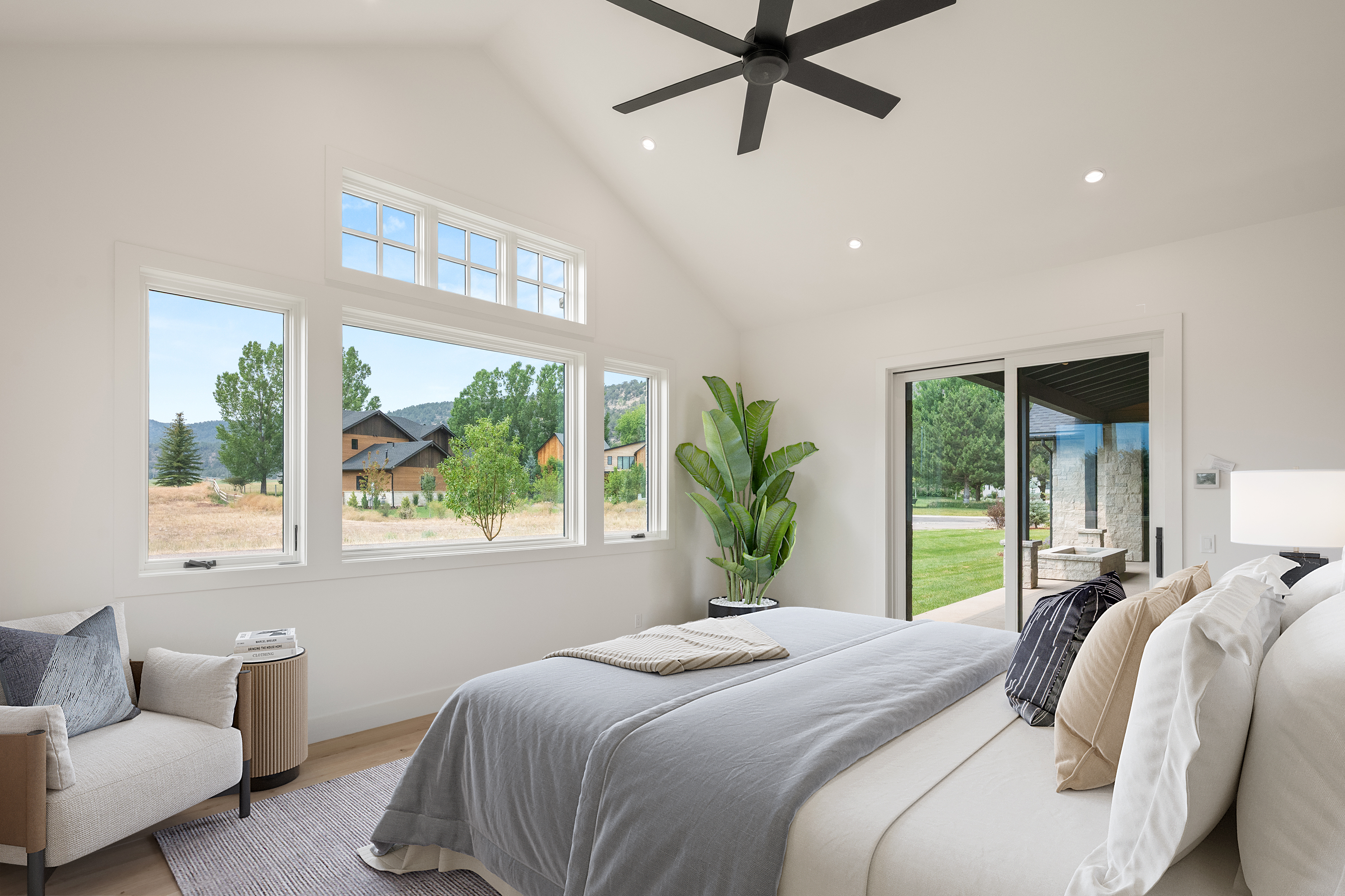 This serene and modern primary bedroom features high vaulted ceilings with a matte black ceiling fan, creating an airy and sophisticated atmosphere. Large windows and a glass sliding door fill the space with natural light while offering scenic outdoor views and access to the patio. Neutral tones, a cozy reading chair, and an elegant bed ensemble combine to provide a luxurious and inviting sanctuary.