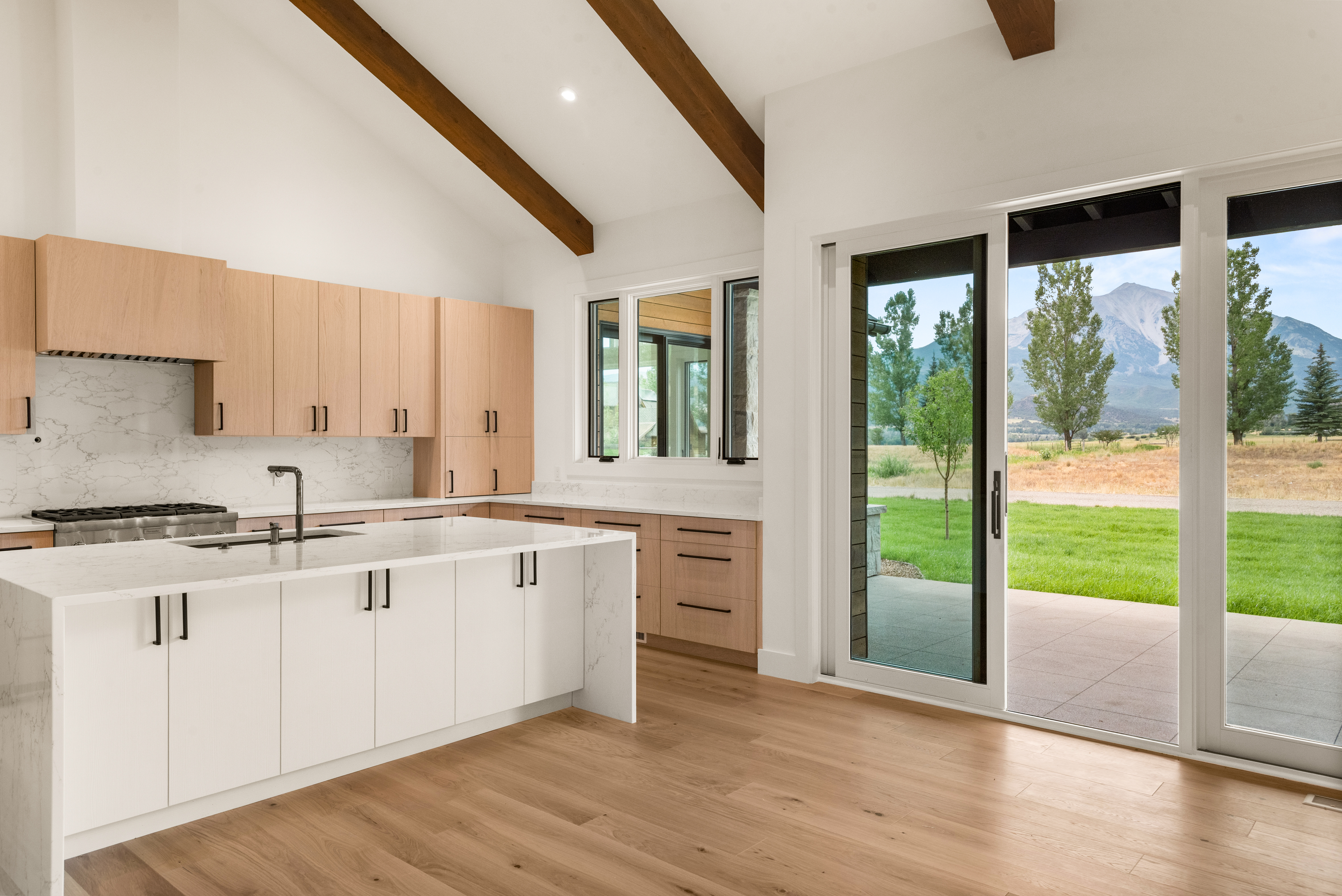 This sleek, modern kitchen features a striking contrast between white kitchen island cabinetry and light wood-tone wall cabinets, complemented by elegant marble-patterned quartz countertops and a matching backsplash. Large sliding glass doors provide a seamless transition to the outdoors, framing a beautiful mountain landscape and expansive yard. The high, vaulted ceiling with dark wood beams adds architectural character and a spacious, airy feel to the home.