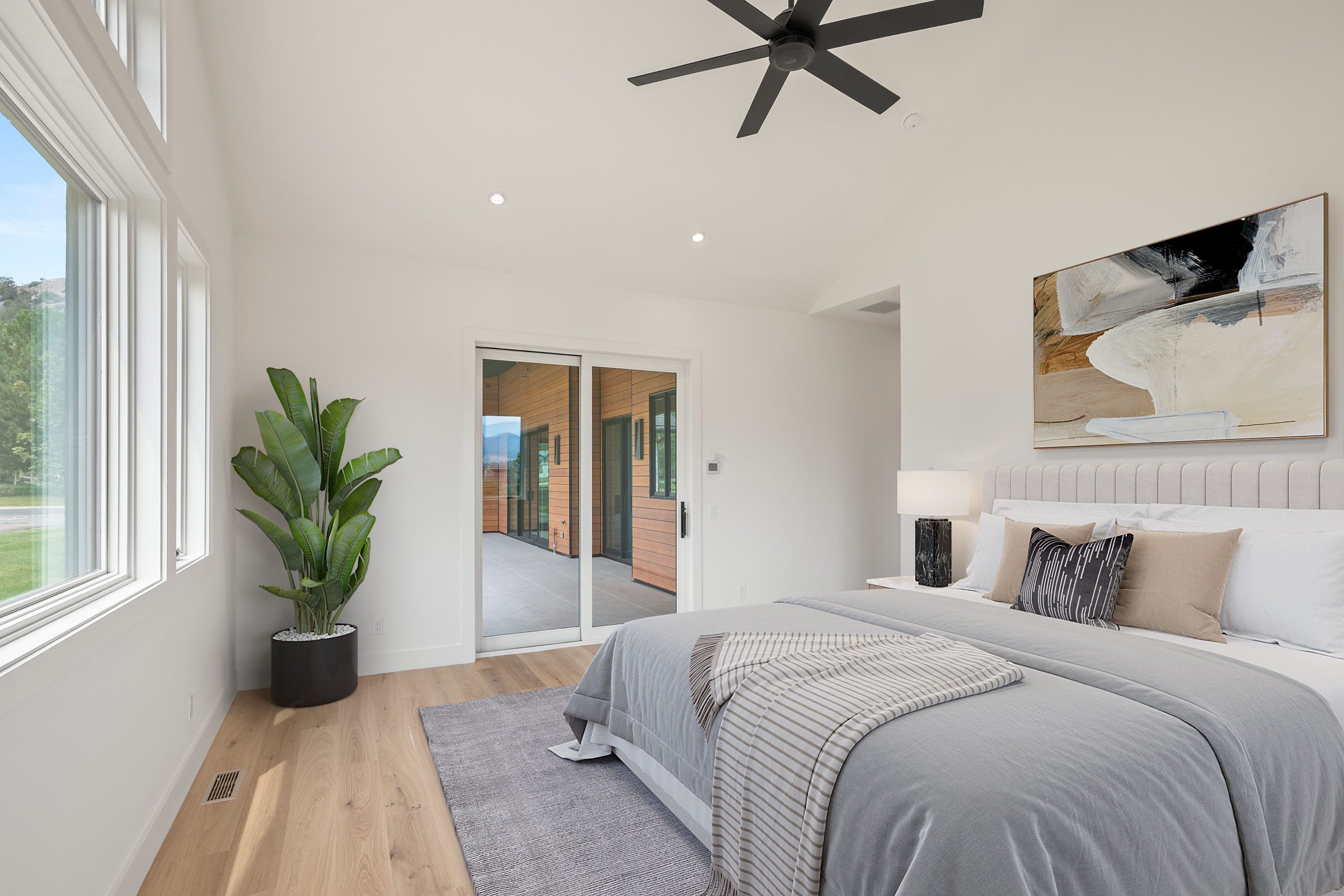 This bright and airy primary bedroom features a modern, neutral aesthetic with light hardwood floors and a soft gray color palette. The room is highlighted by large windows that allow for ample natural light, a sleek sliding glass door leading to an outdoor deck, and contemporary decor, including a textured accent pillow and minimalist lighting. The space offers a calm, sophisticated atmosphere perfect for a master suite.