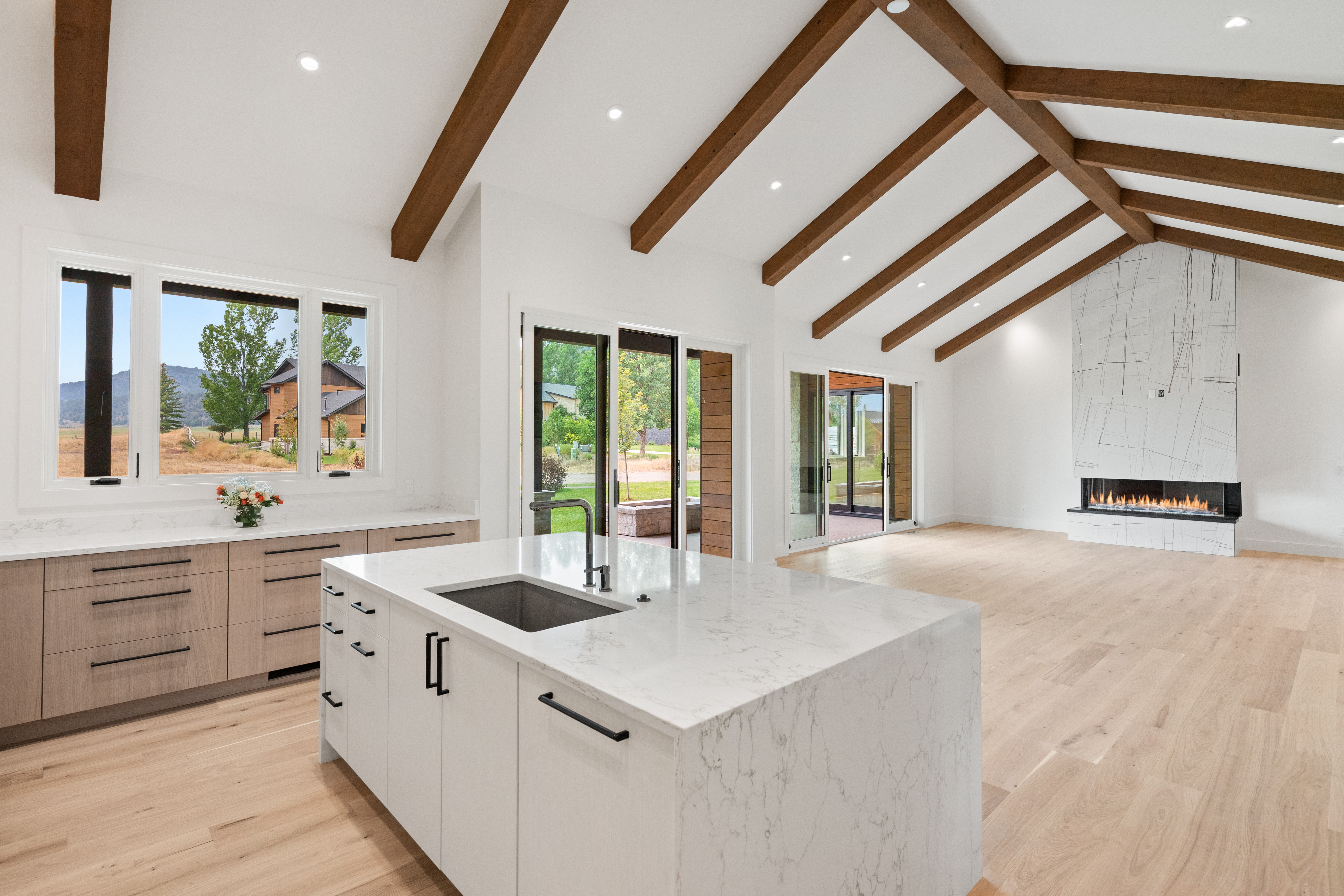 This stunning open-concept kitchen features a modern aesthetic with light wood-tone cabinetry, a central white quartz island, and sleek black hardware. The space is highlighted by vaulted ceilings with exposed wood beams and a contemporary fireplace, creating a bright and airy atmosphere. Large glass sliding doors and windows provide seamless indoor-outdoor flow and beautiful natural light.