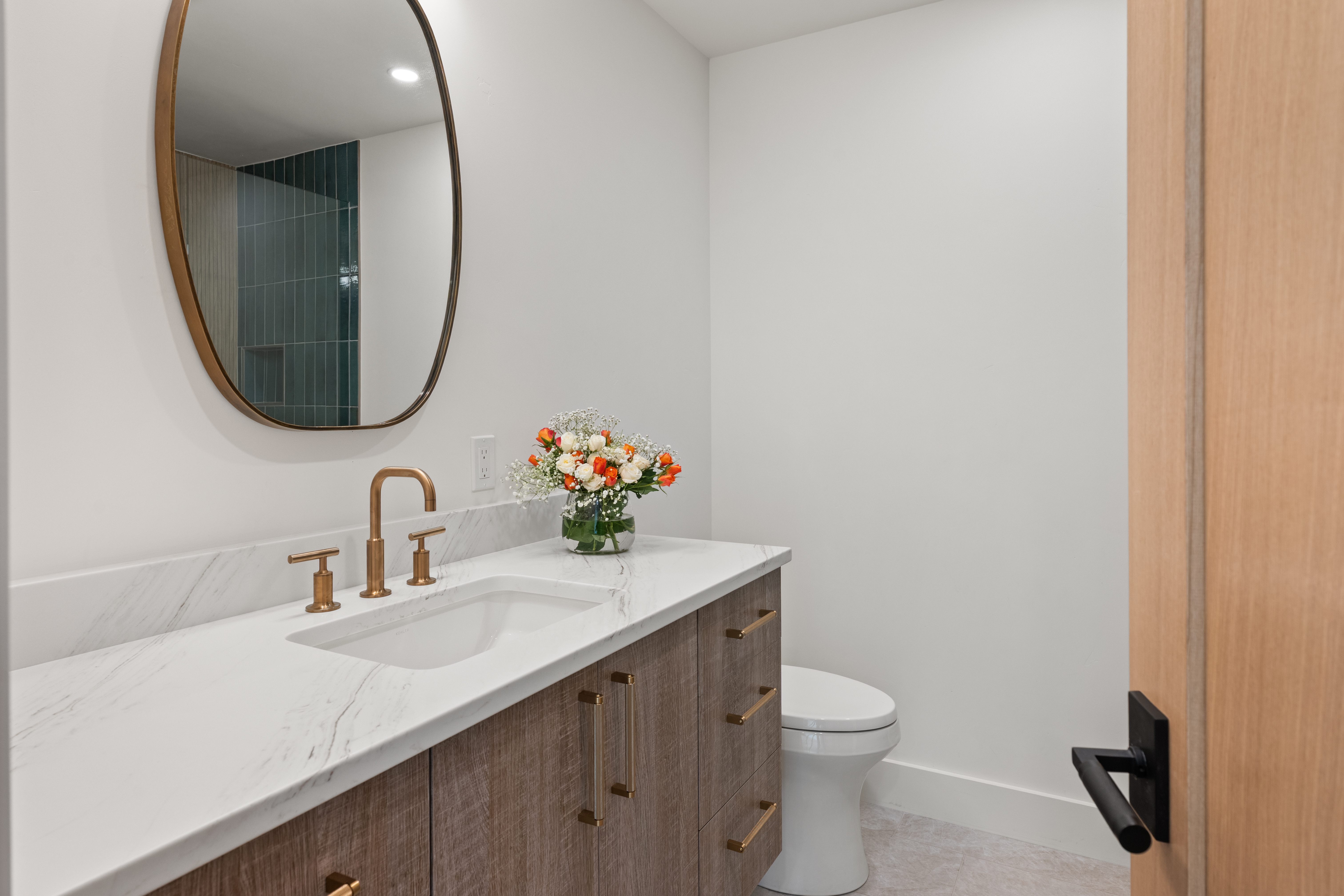 This modern bathroom features a stylish floating vanity with wood-grain cabinetry, a white marble-patterned countertop, and elegant brushed-gold fixtures. An oval mirror hangs above the sink, complementing a clean, minimalist aesthetic highlighted by a small vase of fresh flowers. The space is well-lit and includes a sleek white toilet, presenting a polished and contemporary powder room design.