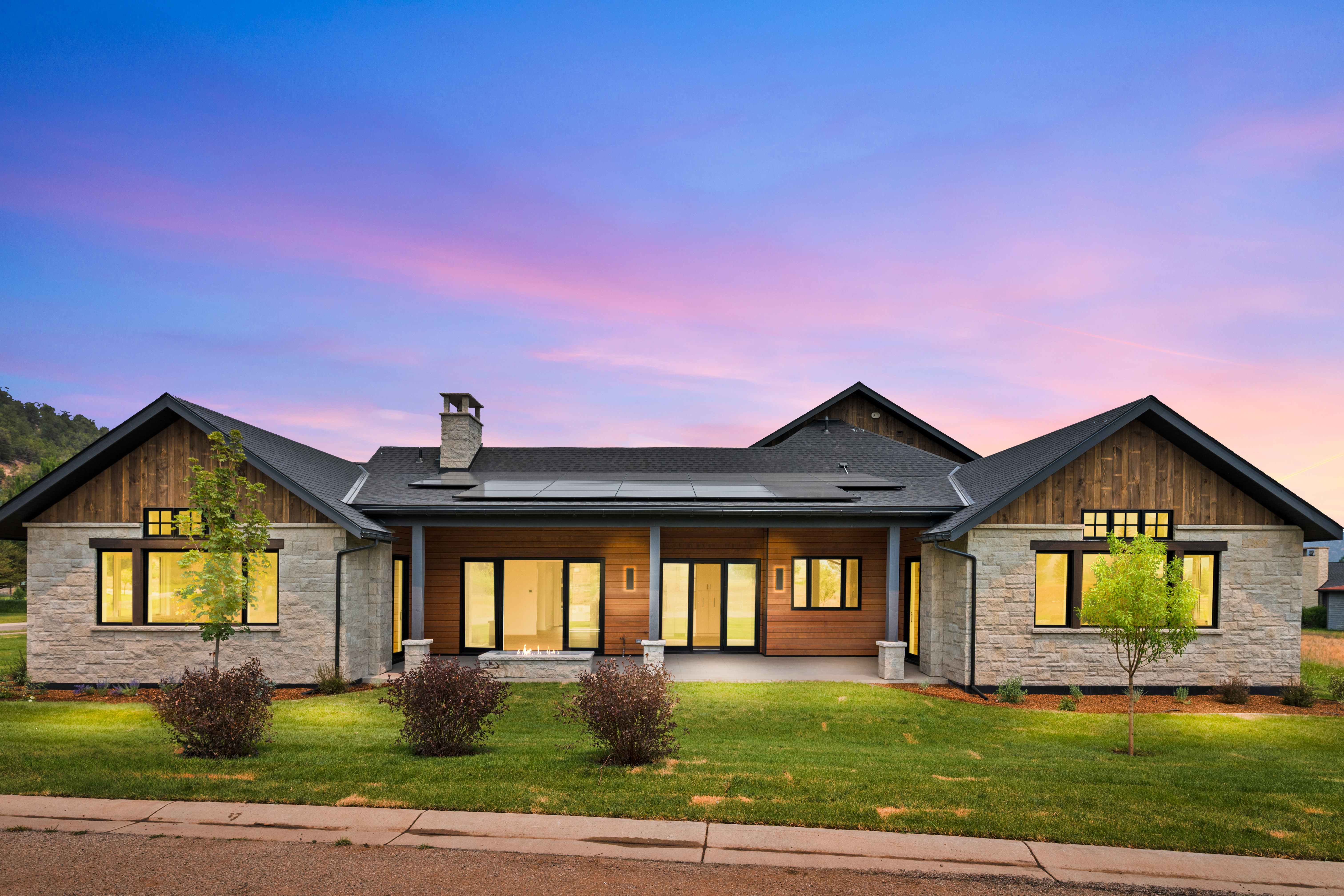 This stunning contemporary residence showcases a sophisticated blend of natural stone and warm wood siding, accented by dark window frames and trim. The home features a prominent covered patio area with an integrated fire pit, perfect for outdoor living, and is topped with high-quality shingles and solar panels. Set against a vibrant twilight sky, the beautifully landscaped lawn and well-placed architectural lighting give the property an inviting and high-end curb appeal.