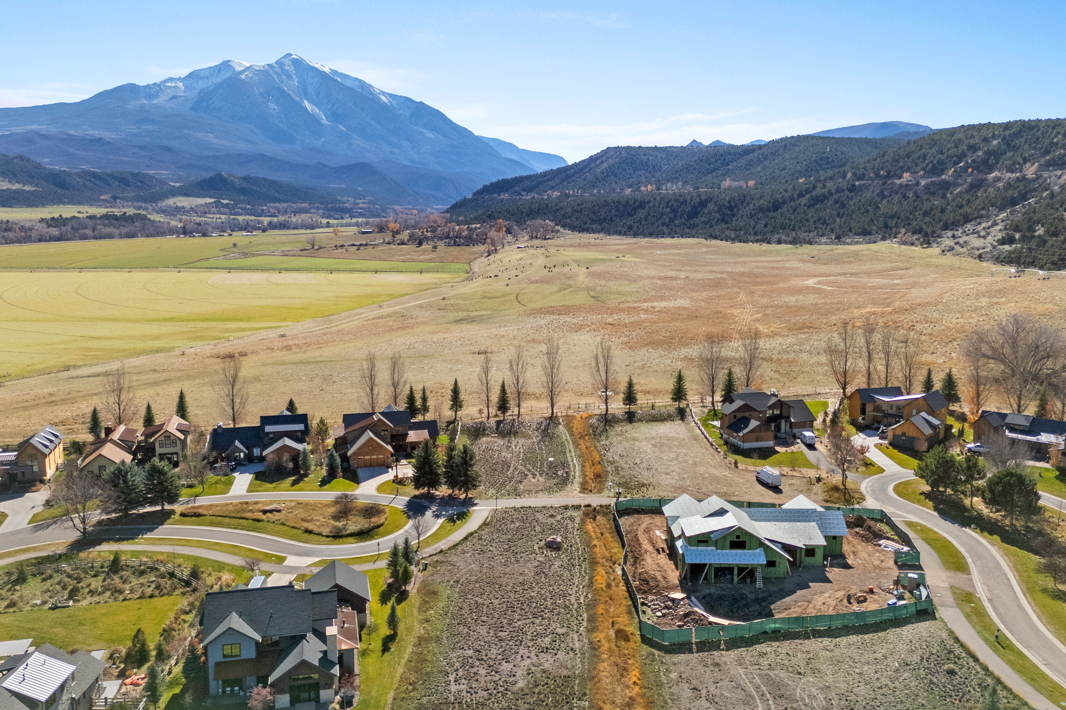 This aerial view captures a luxury residential community set against a breathtaking backdrop of vast, snow-capped mountains and expansive open fields. The neighborhood features high-end homes with architectural styles blending modern and rustic elements, along with a prominent new construction site. The setting offers a serene, spacious environment that highlights both the natural beauty of the landscape and the upscale character of the residential development.