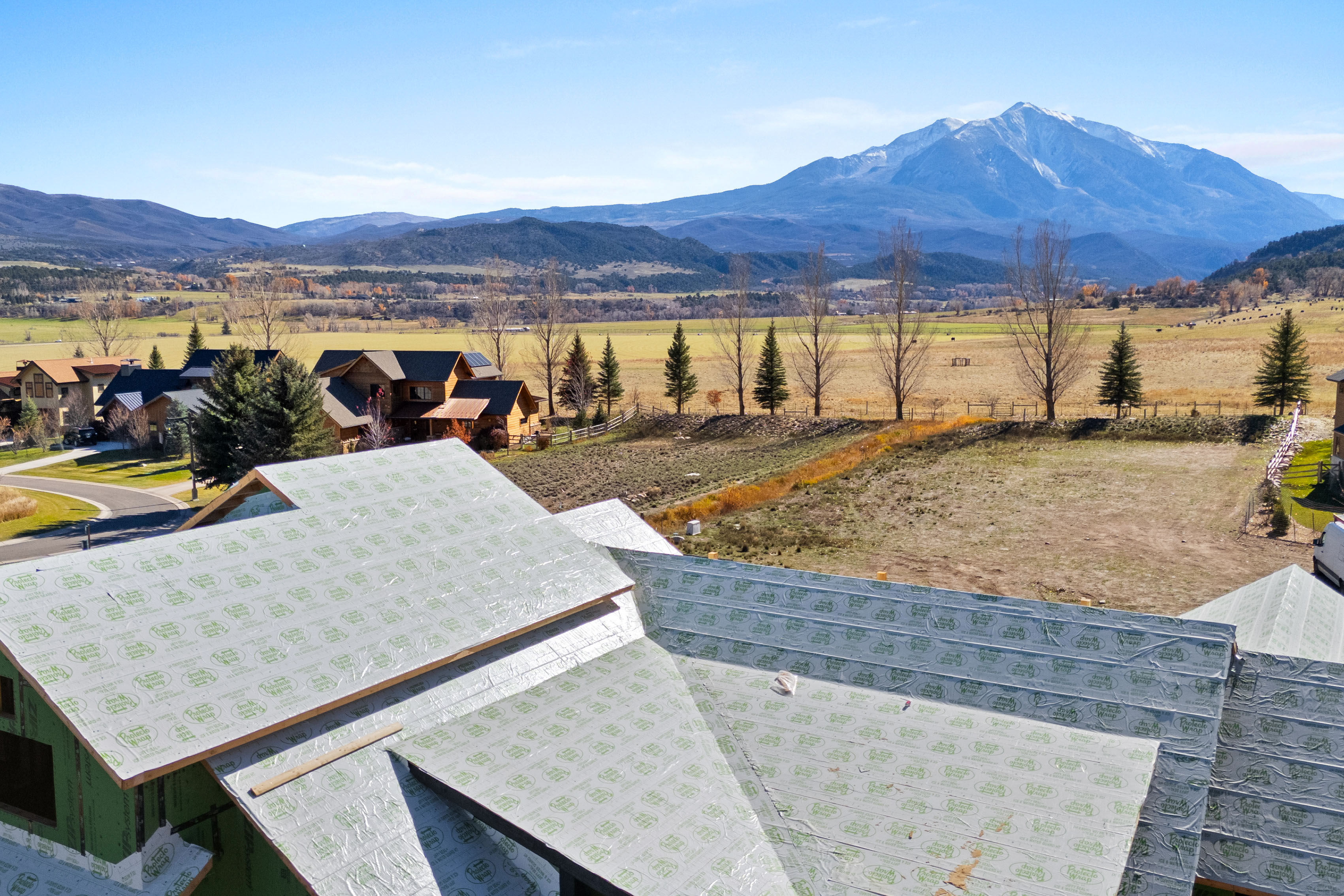 This aerial view showcases a property under construction, highlighting the expansive roof structure covered in protective underlayment. The elevated perspective captures stunning panoramic vistas of the surrounding valley and majestic, snow-capped mountains. The property is situated in a scenic neighborhood, offering a promising combination of new development and natural beauty.