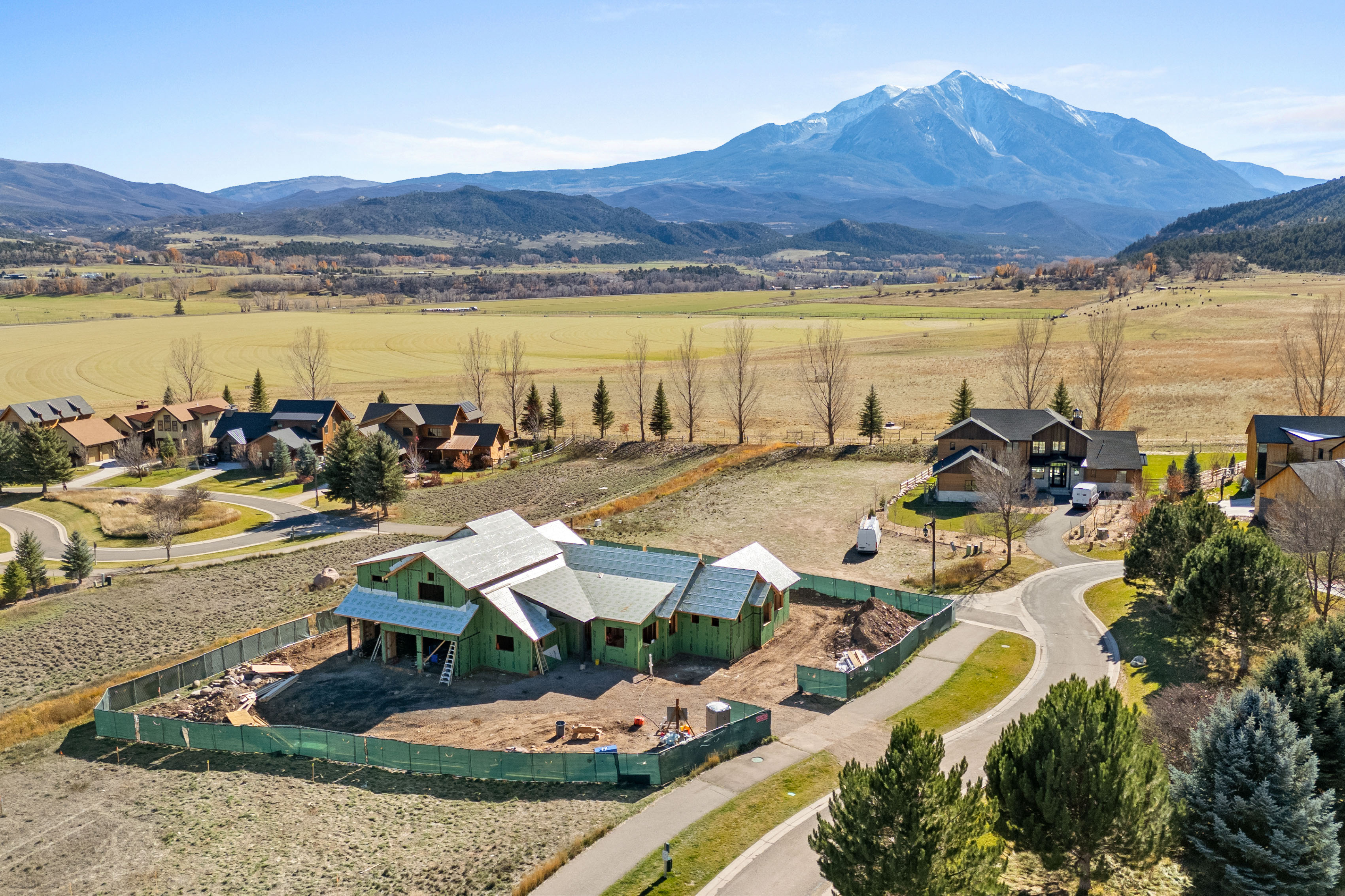 This high-angle aerial view showcases a residential development featuring a new construction project framed in green sheathing, situated among completed luxury homes. The setting offers expansive, scenic views of a vast valley floor and a prominent snow-capped mountain range in the background. The property layout highlights the suburban design within a peaceful, rural-adjacent landscape.