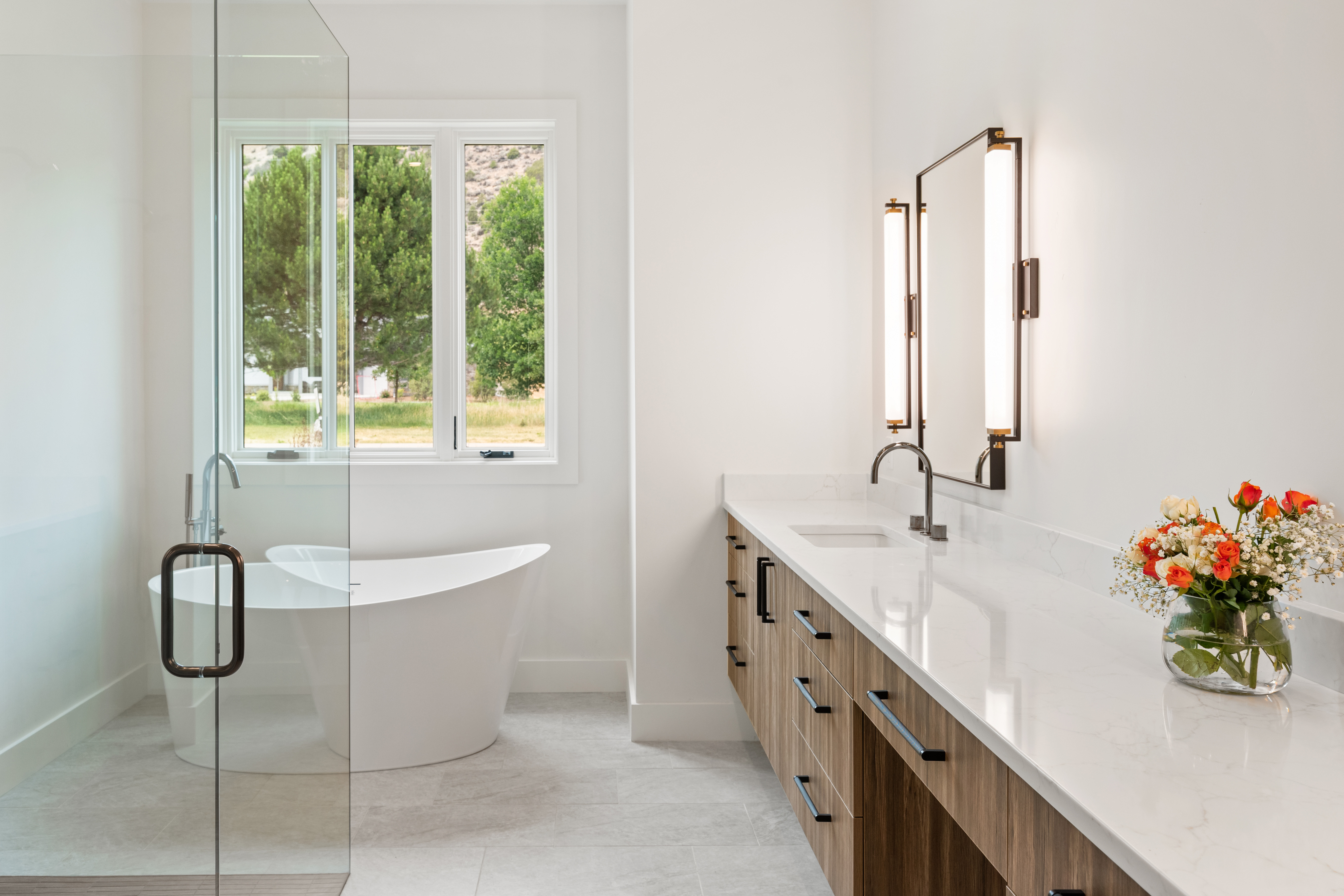 This sleek and modern primary bathroom features a floating wood vanity with clean-lined hardware, paired with a bright white quartz countertop and a contemporary vessel faucet. A large, frameless glass shower enclosure stands adjacent to a sophisticated freestanding soaking tub, both set against a neutral, minimalist aesthetic. Large windows provide natural light and a pleasant outdoor view, creating an overall atmosphere of refined luxury.