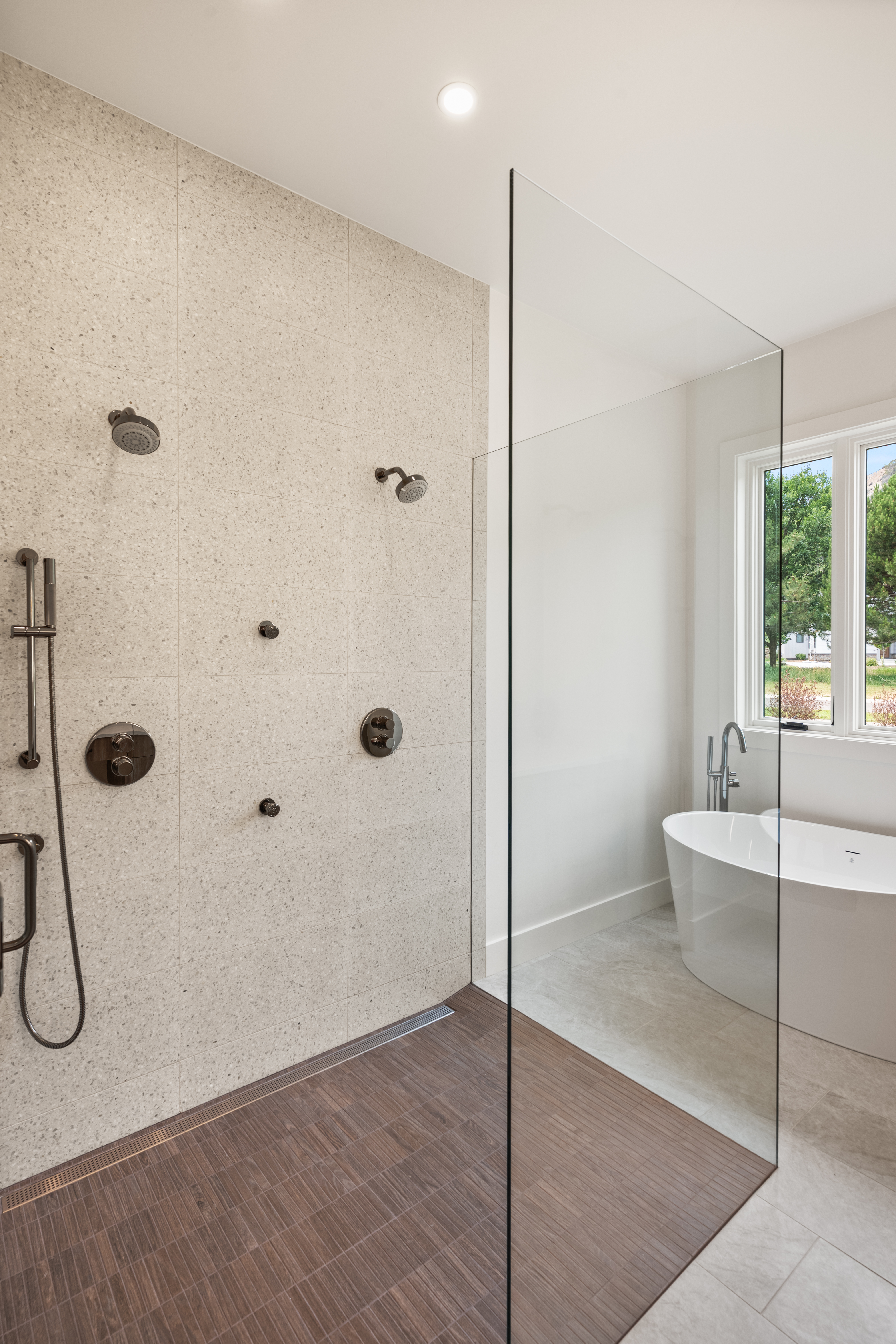 This modern bathroom features a spacious walk-in shower with a sleek glass enclosure and a dark wood-look tile floor that transitions into a bright, airy space. The shower is equipped with multiple fixtures, including a handheld showerhead and wall-mounted sprays, set against neutral speckled wall tiles. A standalone soaking tub is positioned by the window, creating a luxurious and serene spa-like atmosphere.