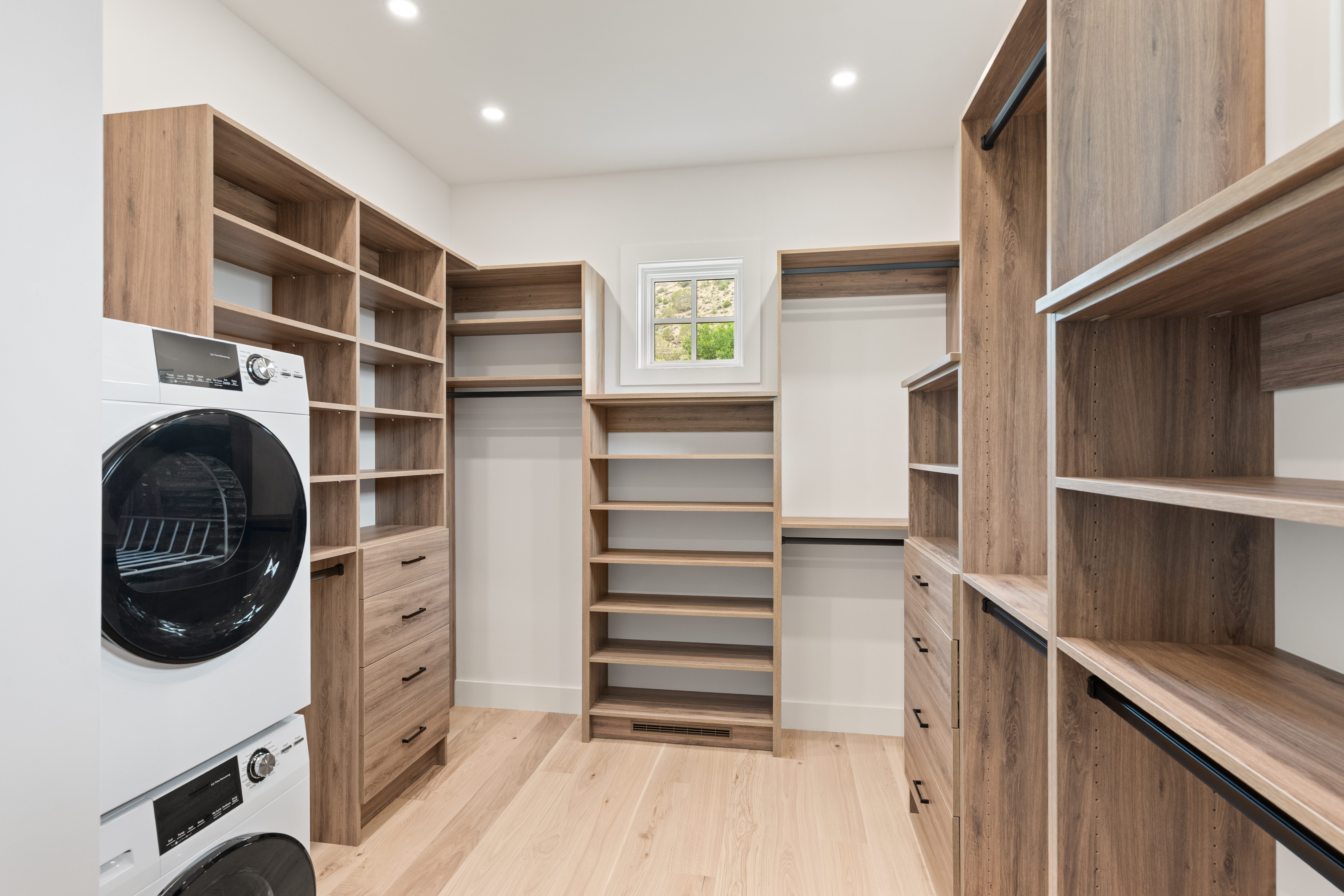 This spacious, modern walk-in closet features custom wood-tone cabinetry, including built-in shelving, hanging rods, and several drawers for ample storage organization. A stackable washer and dryer are seamlessly integrated into the closet layout, adding convenient laundry functionality to the space. The room is completed with light hardwood flooring, bright recessed lighting, and a small window that brings in natural light.