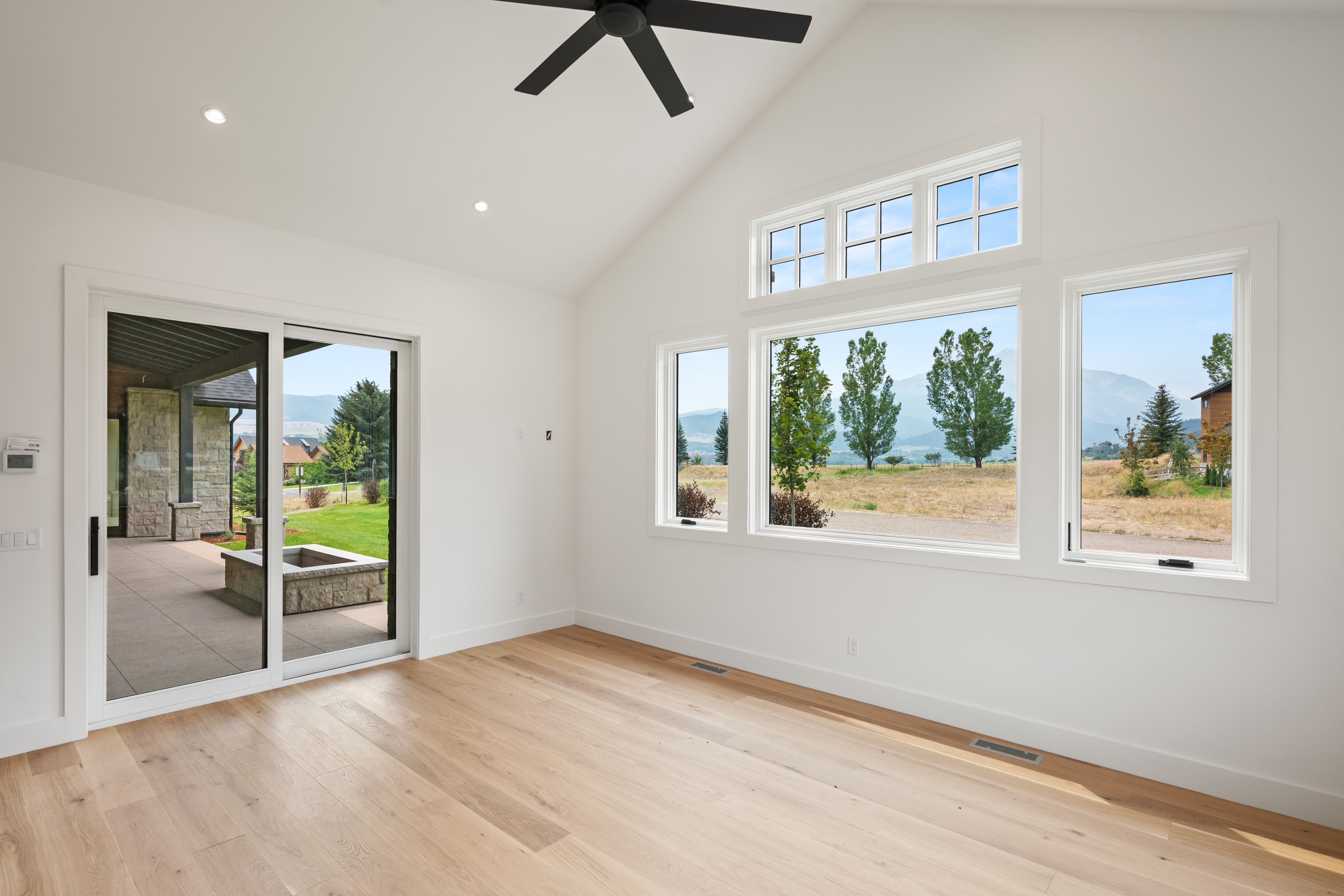 This bright, modern living area features vaulted ceilings, light wood flooring, and a expansive wall of windows that frame picturesque mountain views. A sliding glass door provides seamless access to a covered outdoor patio, enhancing the home's connection to the surrounding landscape. The space is finished in a clean, minimalist style, perfect for contemporary living.