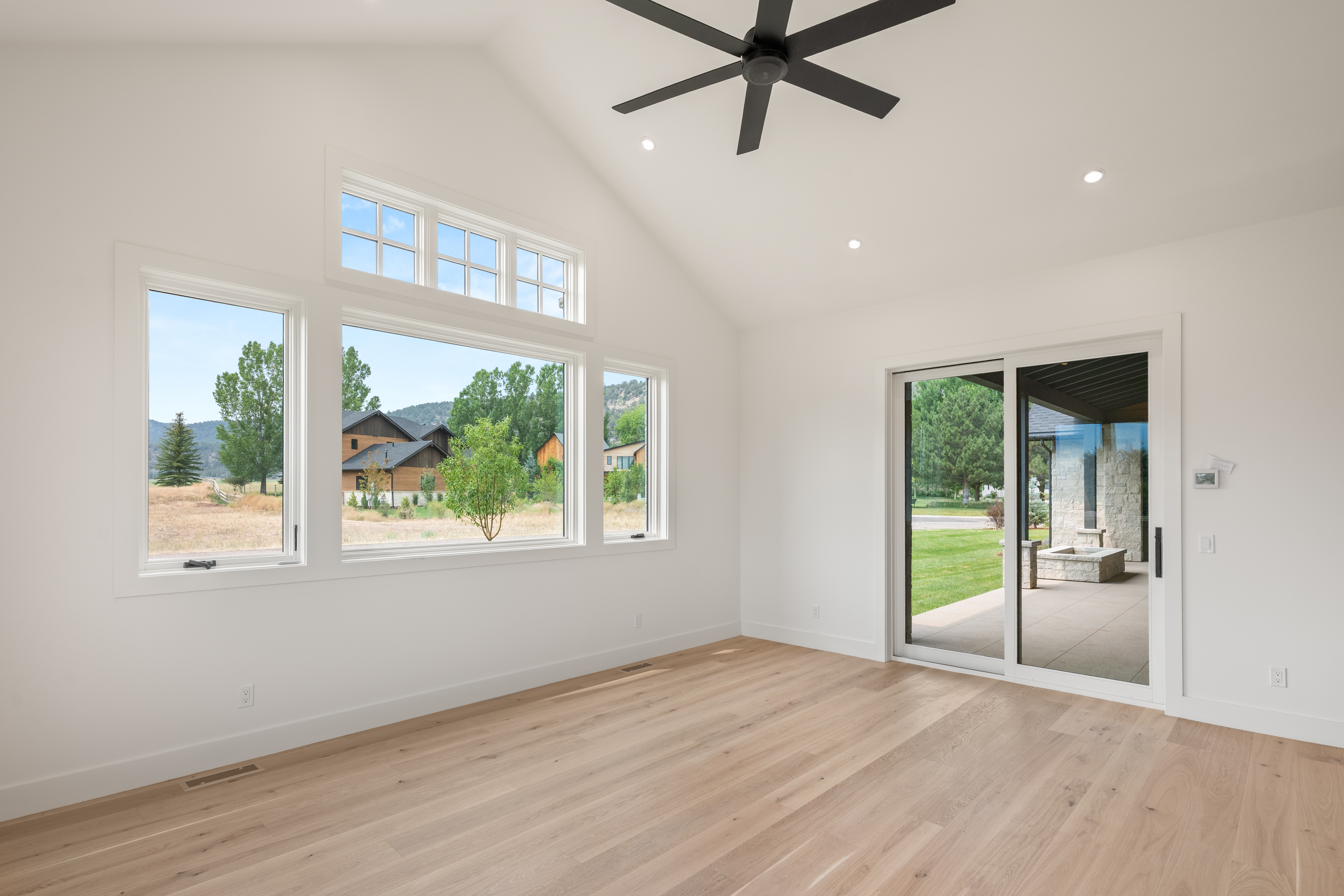 This spacious, light-filled living area features a clean, minimalist aesthetic with vaulted ceilings and beautiful light oak hardwood floors. Large windows and a glass sliding door provide ample natural light and a seamless connection to the outdoor patio and scenic landscape. The room's neutral white palette and modern black ceiling fan create a bright, contemporary atmosphere.