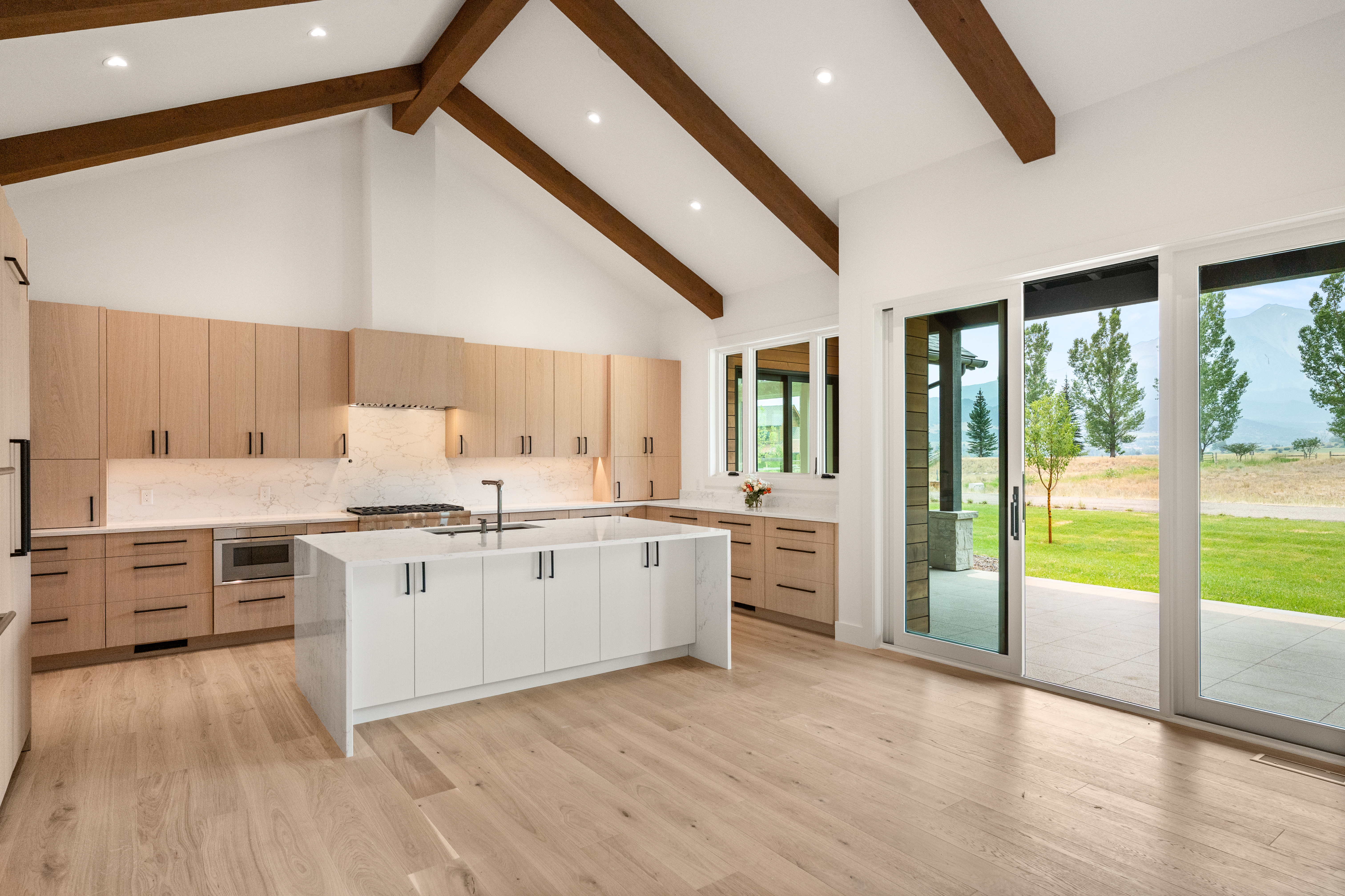 This stunning contemporary kitchen features light wood cabinetry, white marble-veined countertops, and a large central island with seating. The high vaulted ceiling, accented by prominent wood beams, creates an airy and spacious atmosphere, while large sliding glass doors provide seamless indoor-outdoor flow and abundant natural light. The aesthetic is warm and modern, perfectly complemented by the view of the surrounding rural landscape.