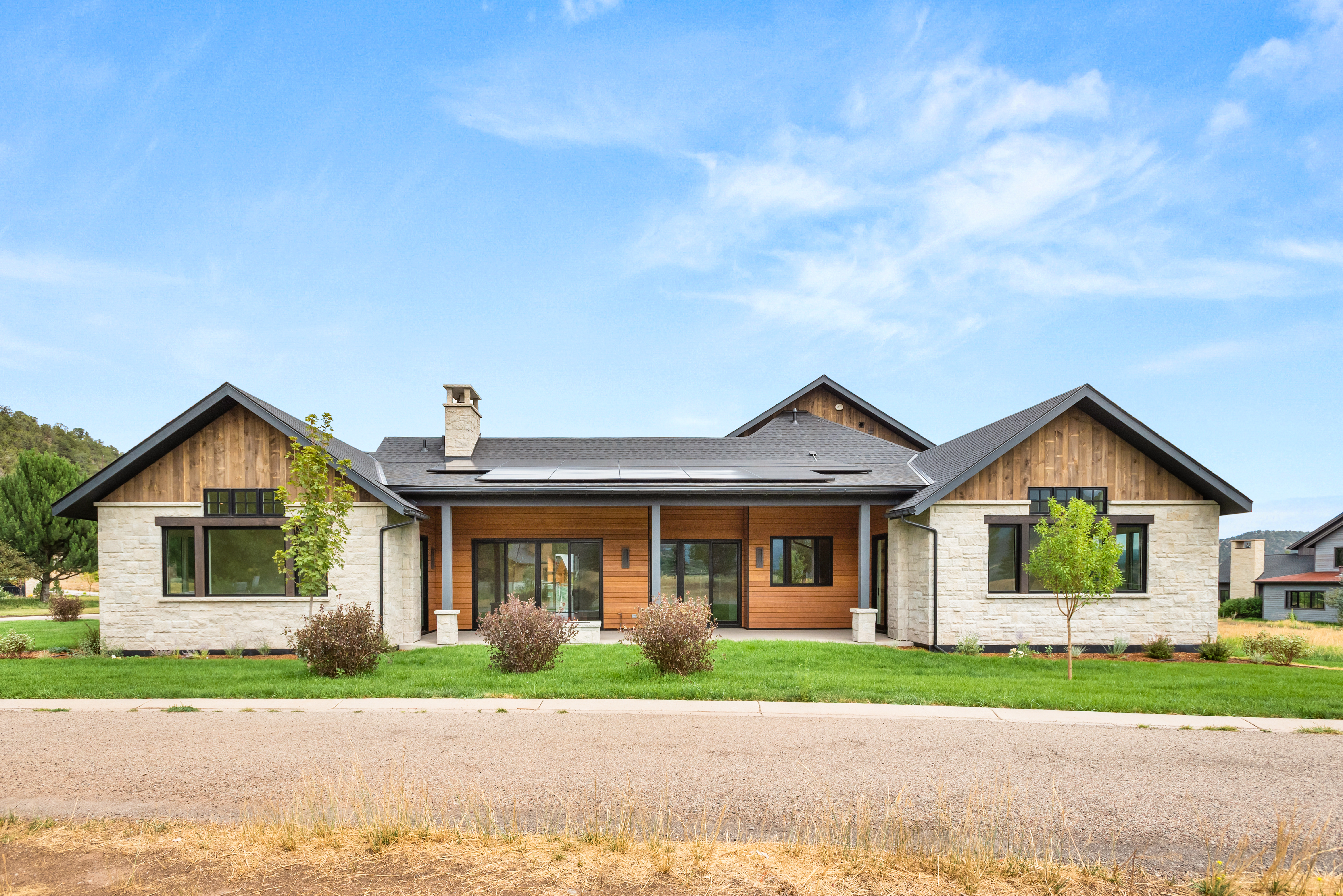 This stunning contemporary residence features a blend of natural stone and warm wood siding, accented by clean architectural lines and a dark metal roof. The exterior includes a spacious covered patio area, large windows that bring in natural light, and solar panels integrated onto the roof. The well-manicured lawn and desert-inspired landscaping frame the home, contributing to its sophisticated and inviting curb appeal.