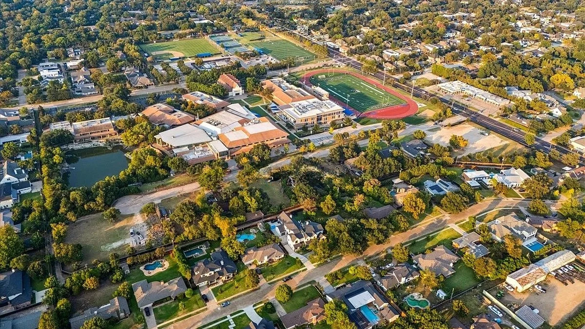 This aerial view captures a well-planned residential area featuring a variety of house styles, lush landscaping, and mature trees. A school or community center stands out prominently, with visible athletic fields and a running track nearby. The scene conveys a sense of established community and neighborhood appeal.