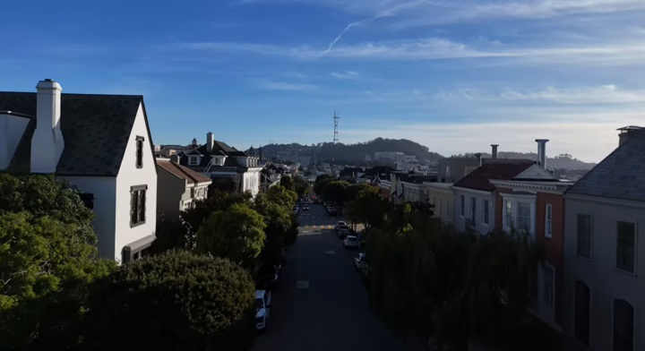 This elevated street view captures a charming residential neighborhood, likely in San Francisco, characterized by historic architectural styles and lined with mature trees. In the distance, Sutro Tower stands prominently against a clear blue sky, serving as a landmark for the locale. The perspective highlights the dense, walkable nature of the community and the well-maintained appeal of the surrounding period homes.