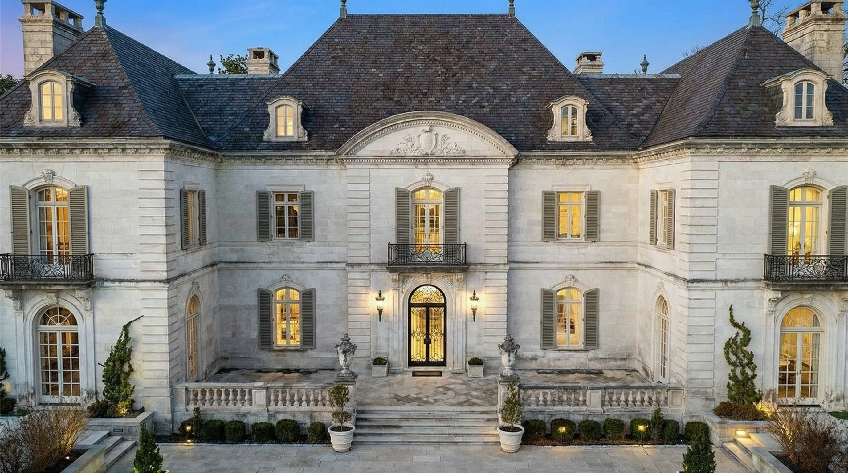This is a front exterior view of a large, luxurious mansion with a symmetrical facade. The building features a light-colored stone exterior, a dark grey roof, and multiple windows with dark shutters. The entryway is grand, with ornate wrought iron doors and decorative lighting, leading to a well-maintained front yard area.