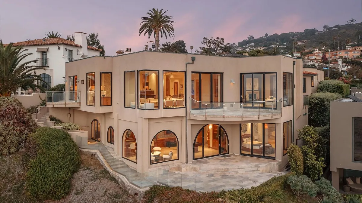 This modern, multi-level luxury residence features a sophisticated beige stucco facade with expansive windows and arched entryways. The home highlights a seamless indoor-outdoor flow, complete with a glass-railed balcony and a spacious, tiered stone patio overlooking the lush hillside. Its contemporary Mediterranean design and prime positioning offer an elegant and airy aesthetic perfect for upscale coastal living.