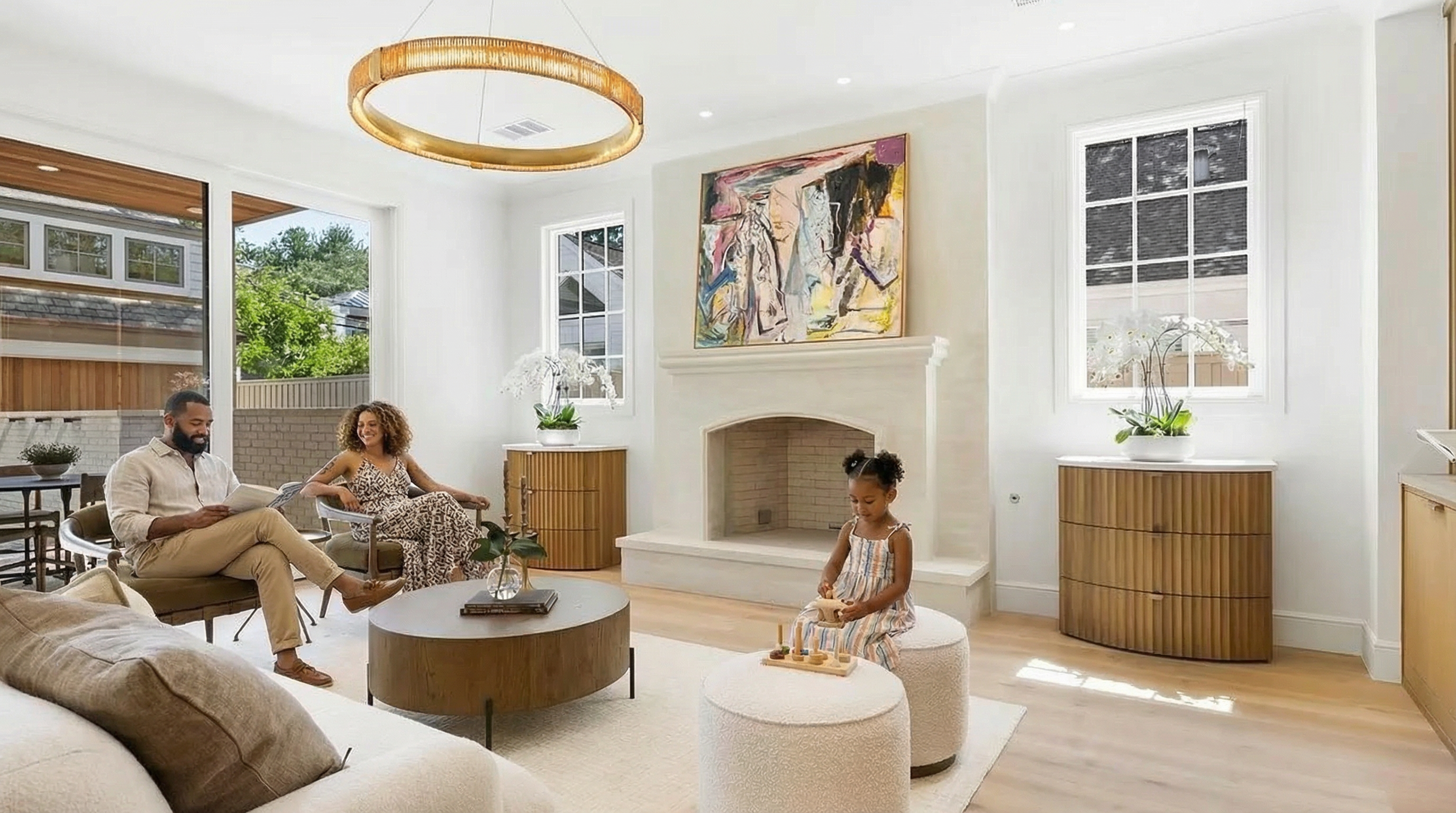 This is a bright and airy living room featuring a contemporary fireplace with a large abstract painting above it. The room is well-lit with natural light from large windows and complemented by a modern circular chandelier. The furniture includes a mix of textures, such as a plush white rug and comfortable seating, offering a welcoming and stylish space for relaxation and entertainment.