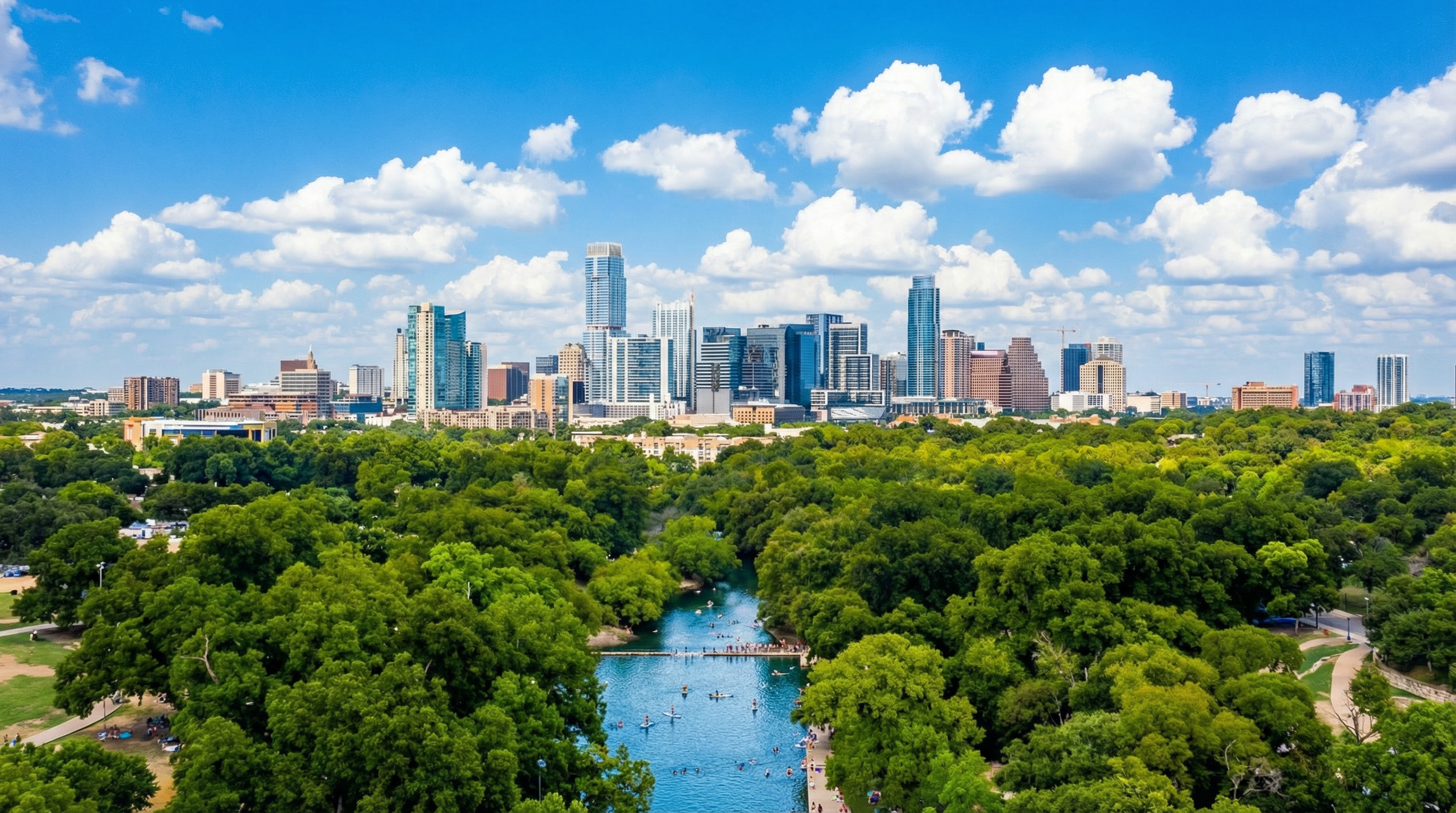This aerial view showcases a vibrant cityscape with modern high-rise buildings set against a backdrop of clear blue skies and fluffy white clouds. Lush green trees line the river running through the city, creating a beautiful contrast between the urban landscape and natural elements. The image suggests a desirable location with access to both city amenities and outdoor recreation.