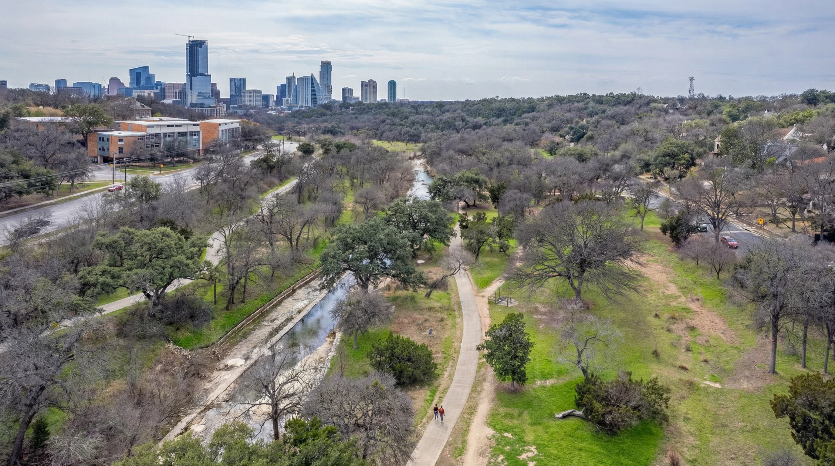 This aerial shot showcases an urban park area with a walking trail and stream running through it, set against the backdrop of a city skyline. The park-like setting offers potential recreational opportunities and adds to the appeal of properties located nearby. This view highlights the proximity to green spaces and city amenities, appealing to those seeking an urban lifestyle with access to nature.